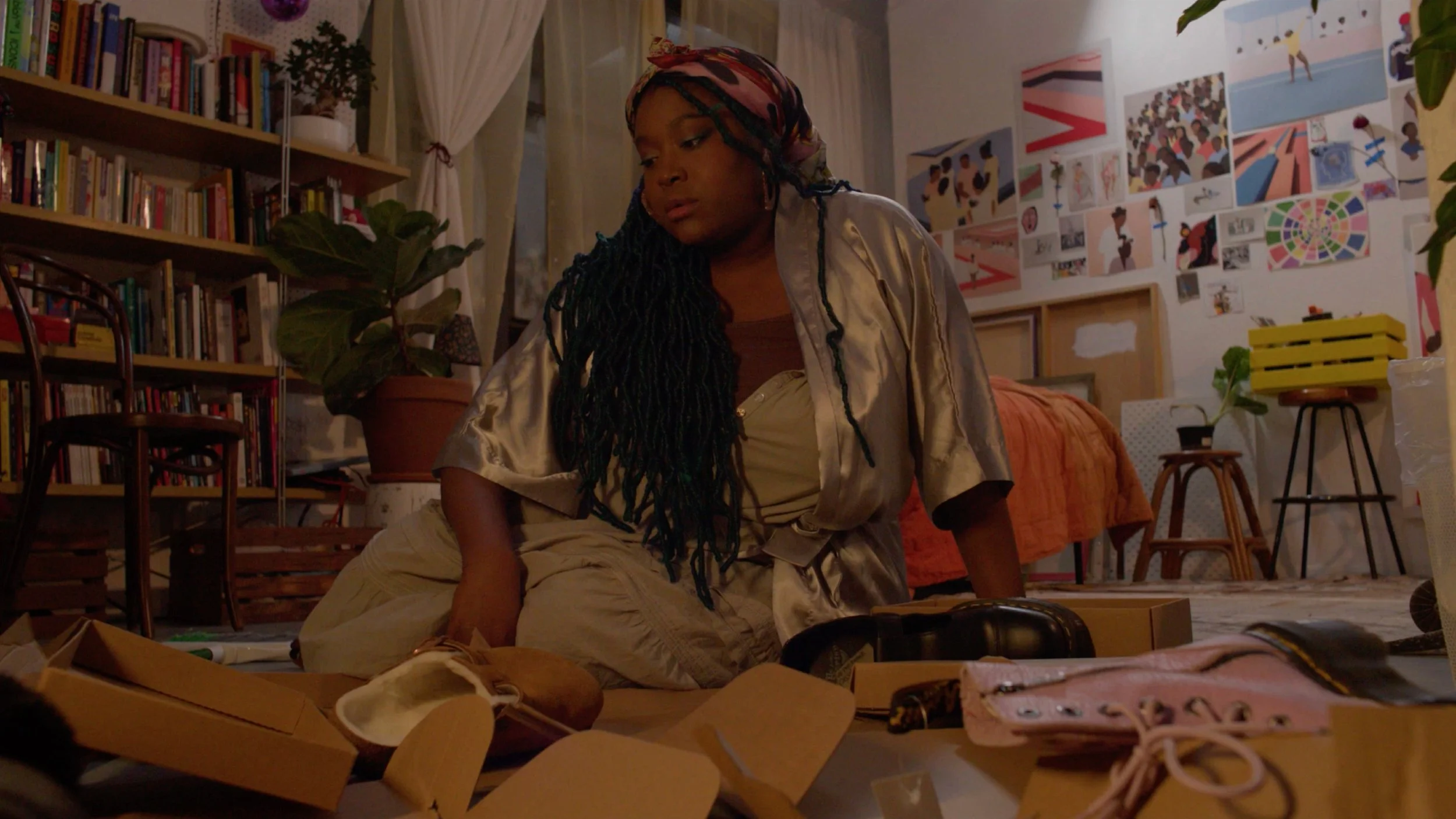 A woman with long dreadlocks and wearing a silk shirt sitting on the floor among scattered boxes and art supplies in a room with colorful artwork on the walls.
