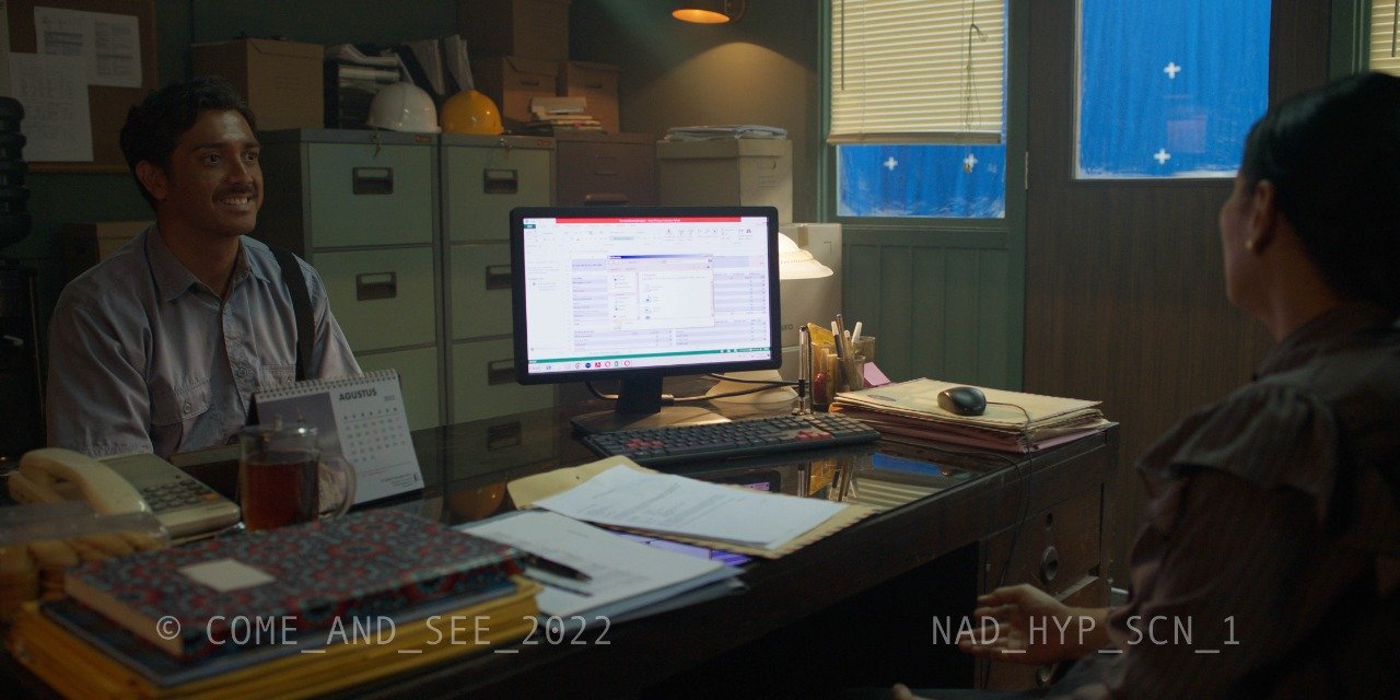 An office with two people, one man and one woman, sitting across from each other. The desk has papers, a telephone, a glass of tea, a computer monitor, and other office supplies. The man is smiling and the woman is facing away from the camera.