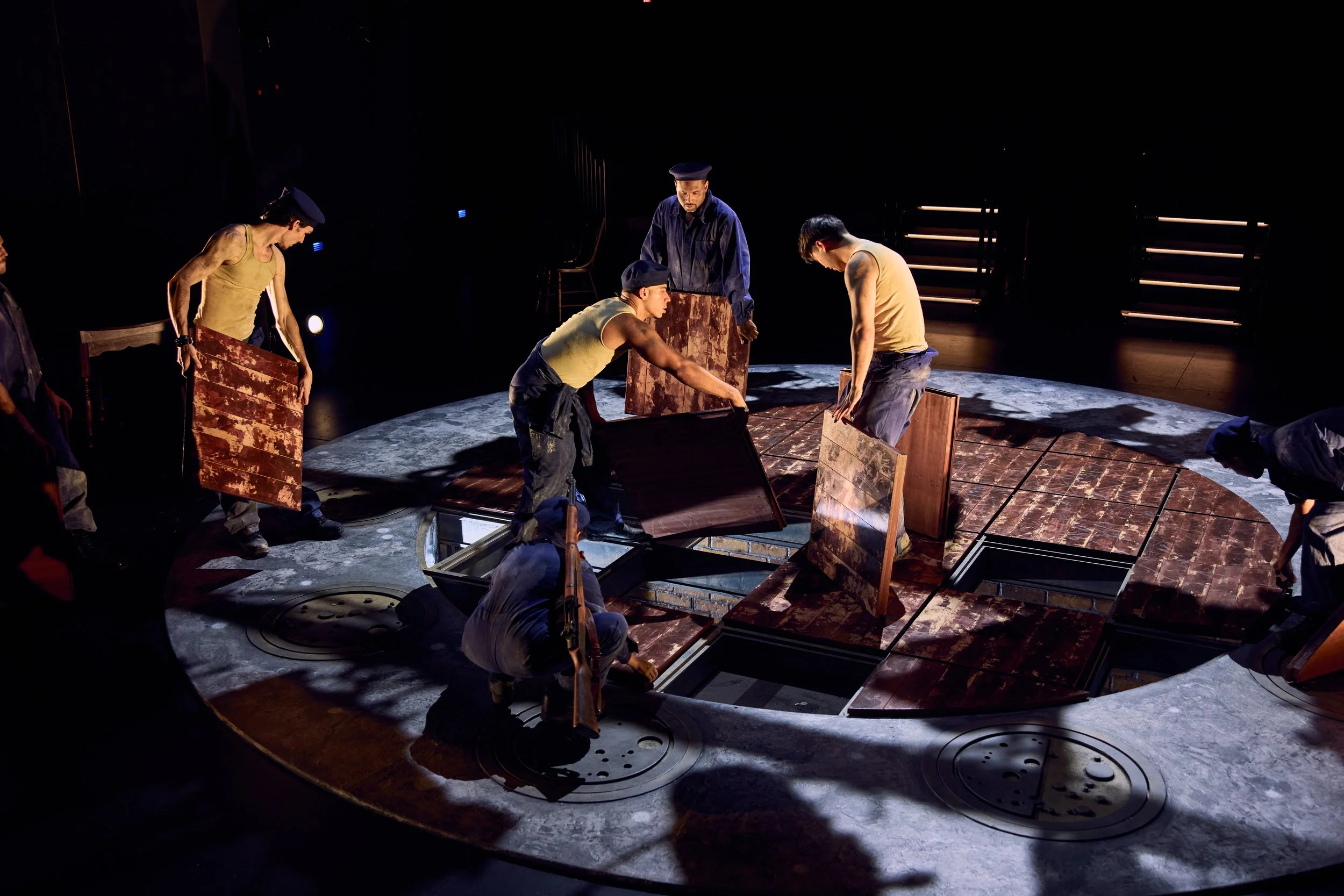 A stage with actors performing a scene involving several wooden panels, with dim lighting focused on the actors, creating a dramatic atmosphere.