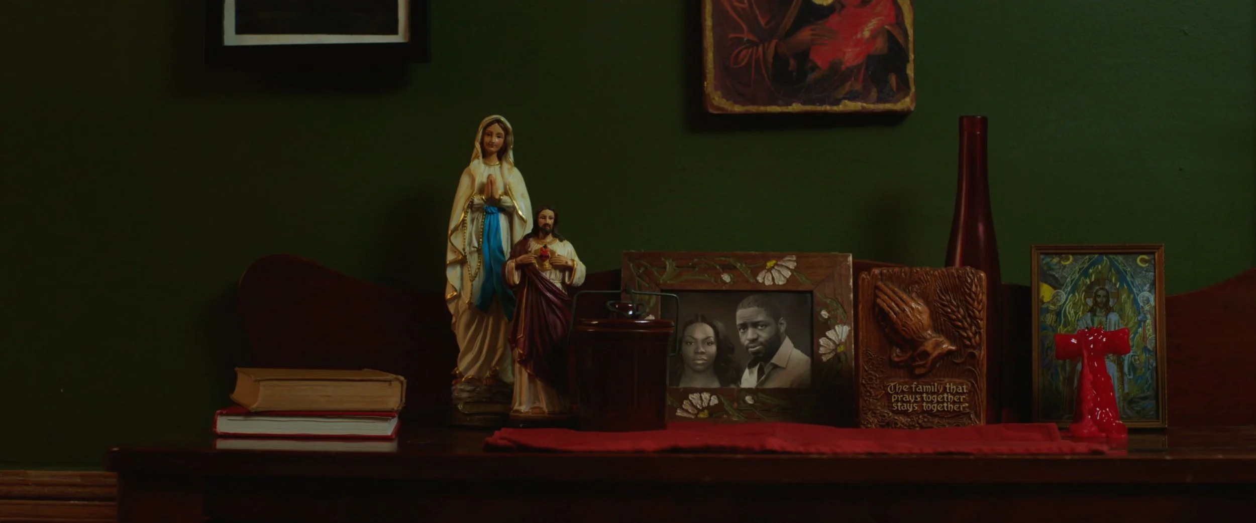 A wooden table with religious statues, framed photographs, and decorative items against a dark green wall. Items include a statue of the Virgin Mary, a framed photo of a couple, a carved wooden plaque with a bird and the phrase 'The family that prays
