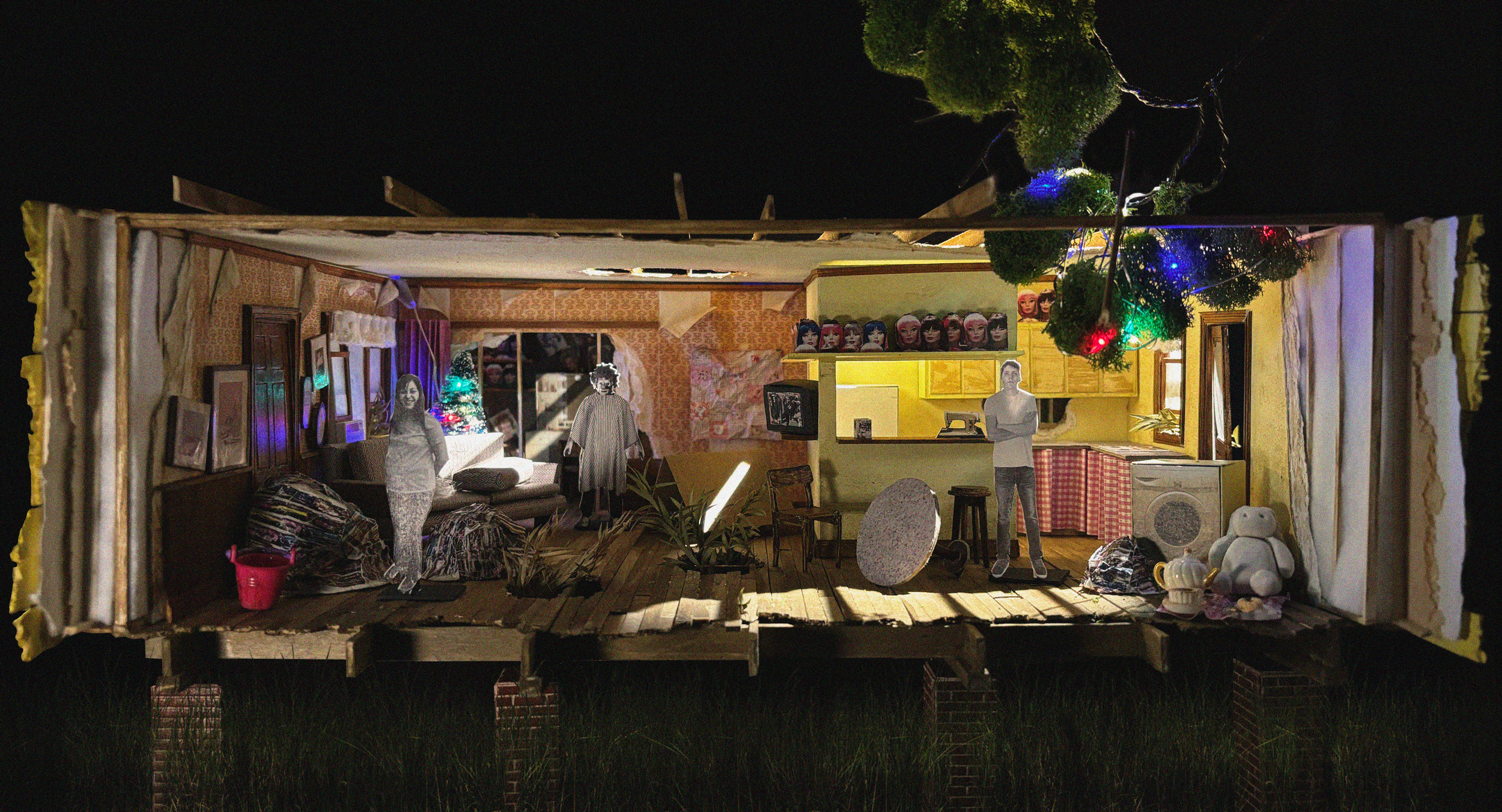 A miniature, dollhouse-style interior of a living room decorated for Christmas at night, with a Christmas tree, string lights, and various furniture and decor items, with three cardboard cutout figures inside.