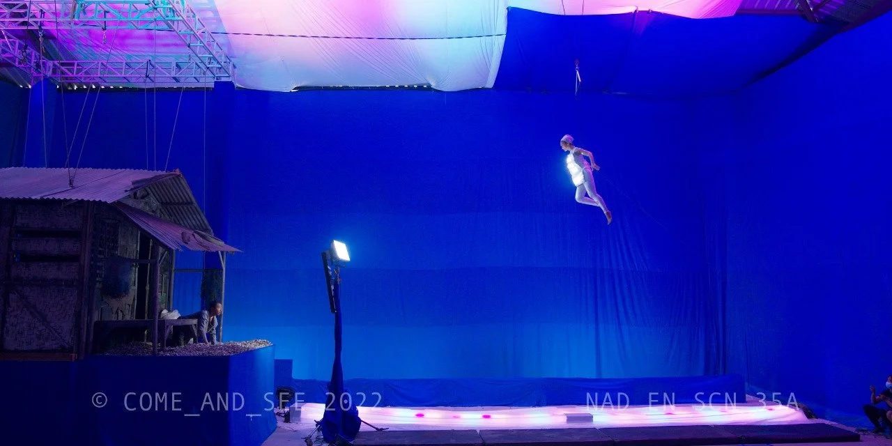 Aerial performer suspended in the air at a circus, illuminated by stage lights, with a backdrop of blue curtains and a small wooden house prop on the stage.