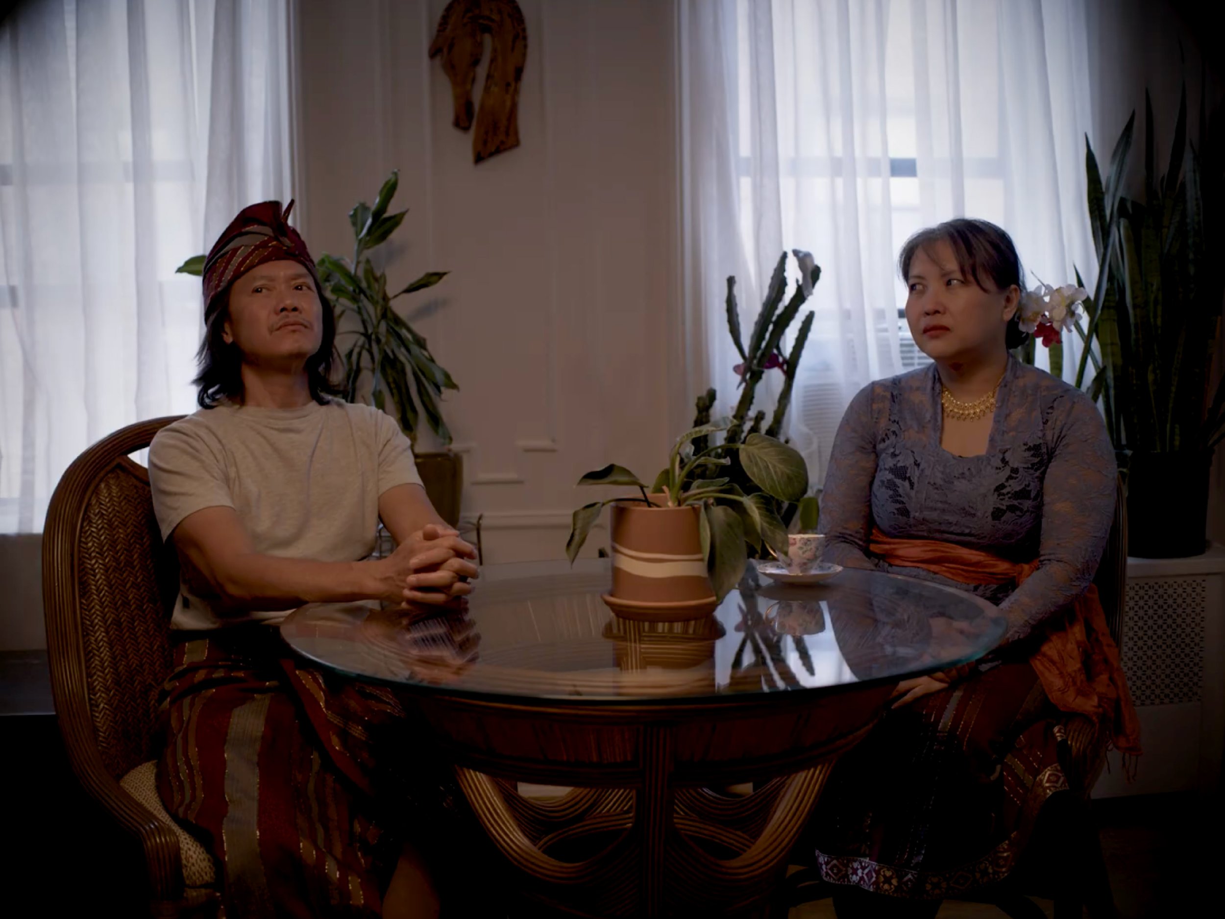 Two people sit at a dining table with houseplants and a decorative wall hanging in the background. The person on the left wears a traditional headscarf and beige shirt, while the person on the right wears a patterned dress and gold jewelry. They appe