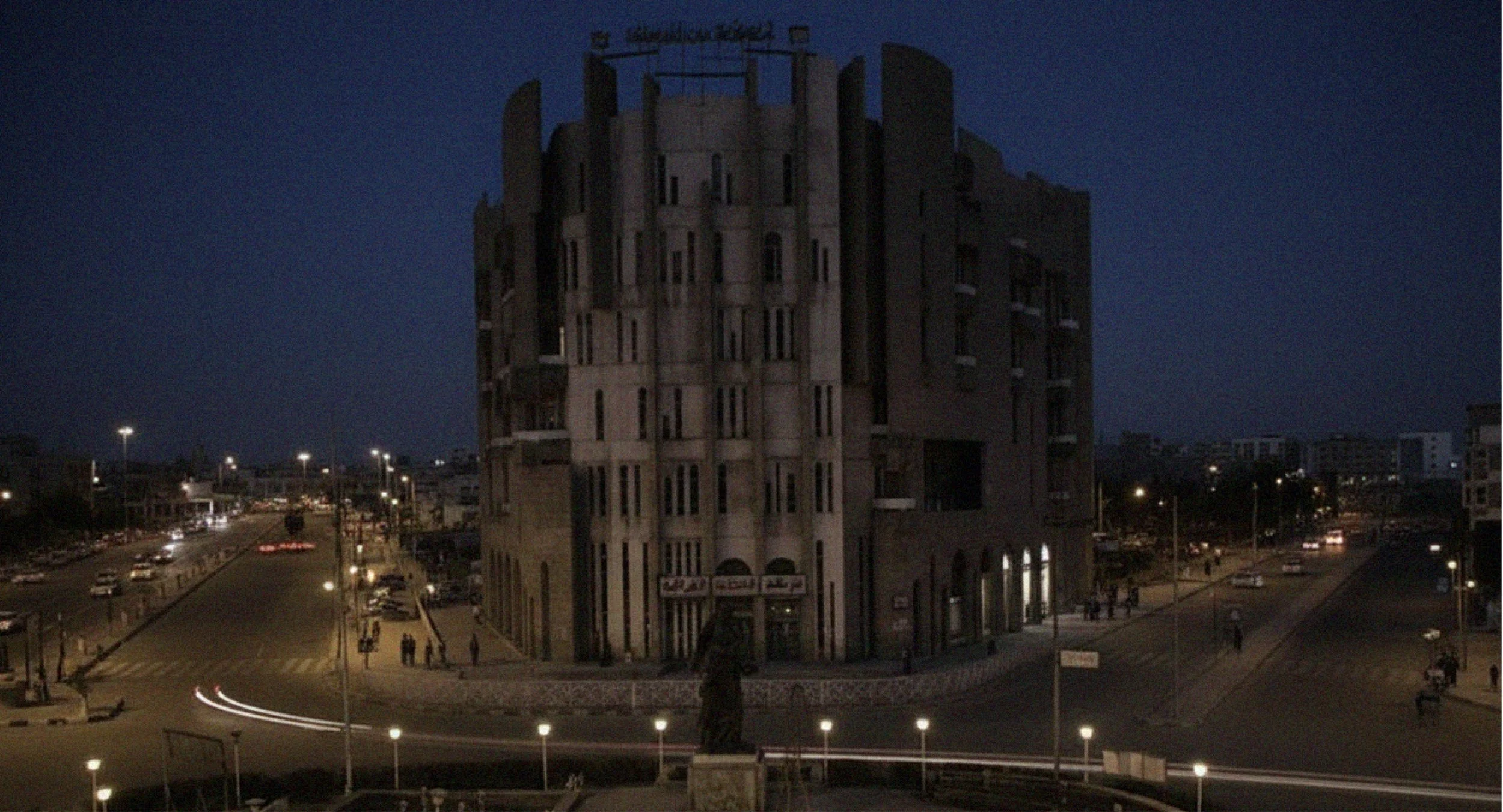 Night view of a tall, historic building with a rounded tower-like structure, illuminated by streetlights, with a busy street and cars below.