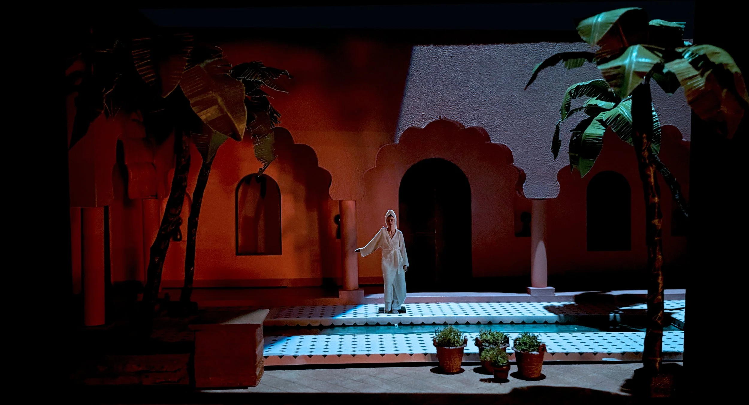 Miniature scene of a woman in a white dress standing on a patterned tile floor, surrounded by stylized palm trees and architectural elements with arched openings and textured walls, illuminated by soft lighting.