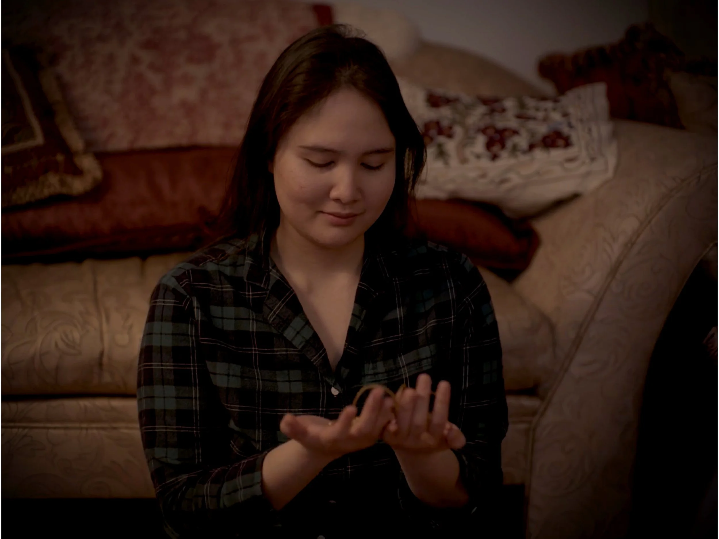 A young woman with brown hair, wearing a black and gray plaid shirt, sitting on a patterned sofa, looking down at a phone in her hands with a slight smile.