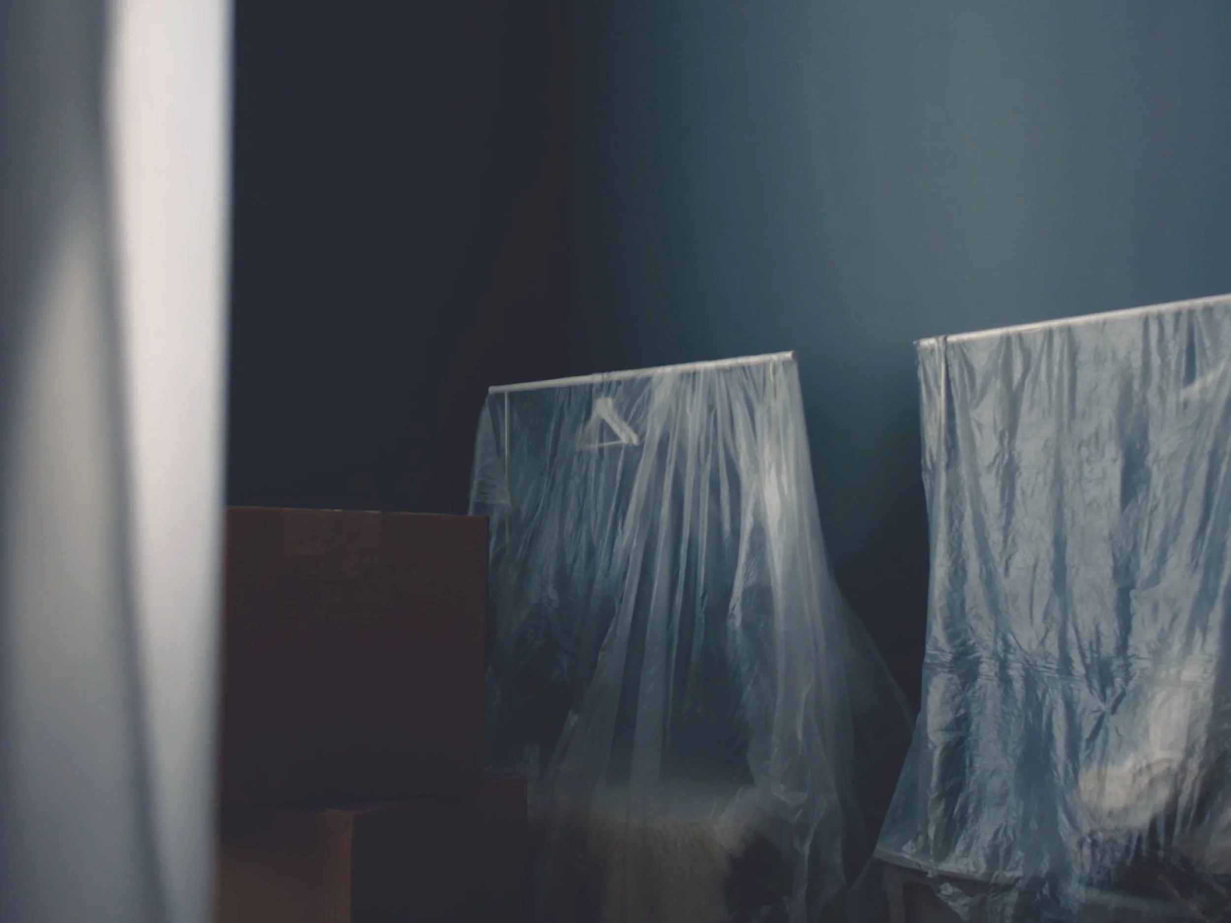 A close-up of a window with plastic covering, partially obscuring a view of a dark interior room with objects covered in plastic or paper.