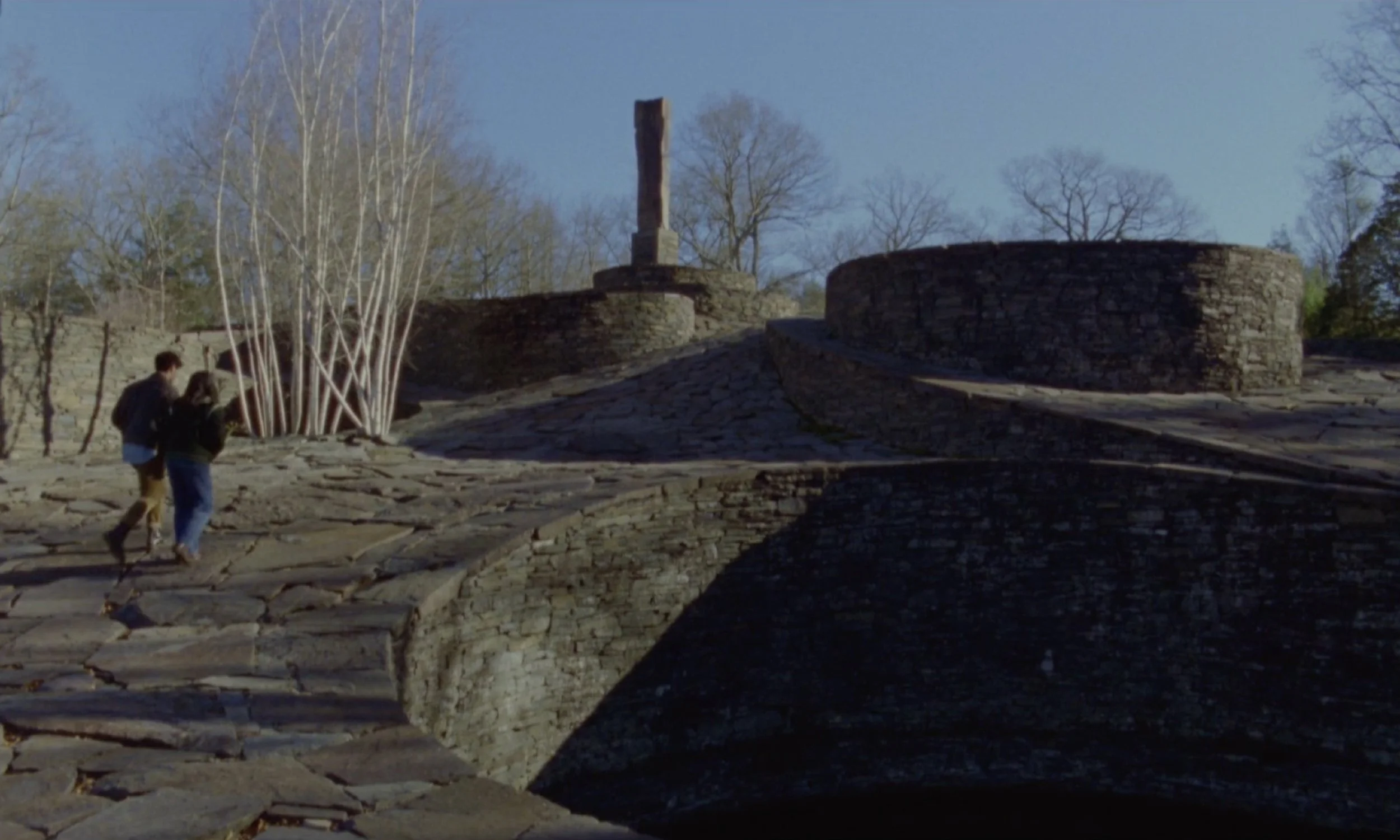 Two people walking along a stone pathway near a stone wall and ruins with tall leafless trees in the background.