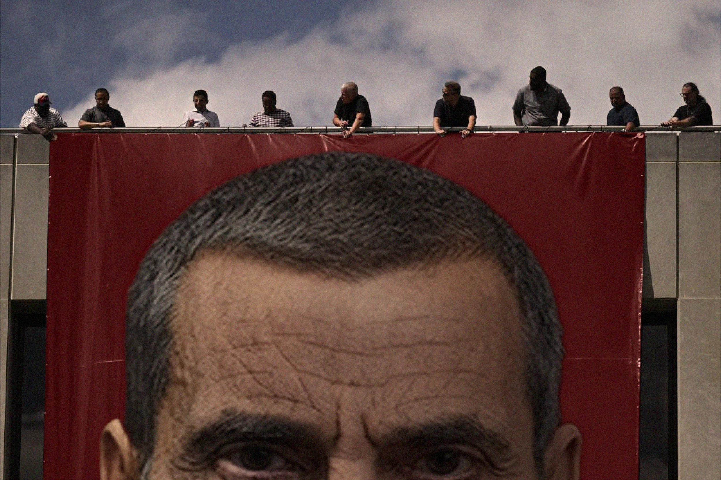A person with gray hair leaning over a red barrier, with several individuals standing along the barrier, against a cloudy sky background.