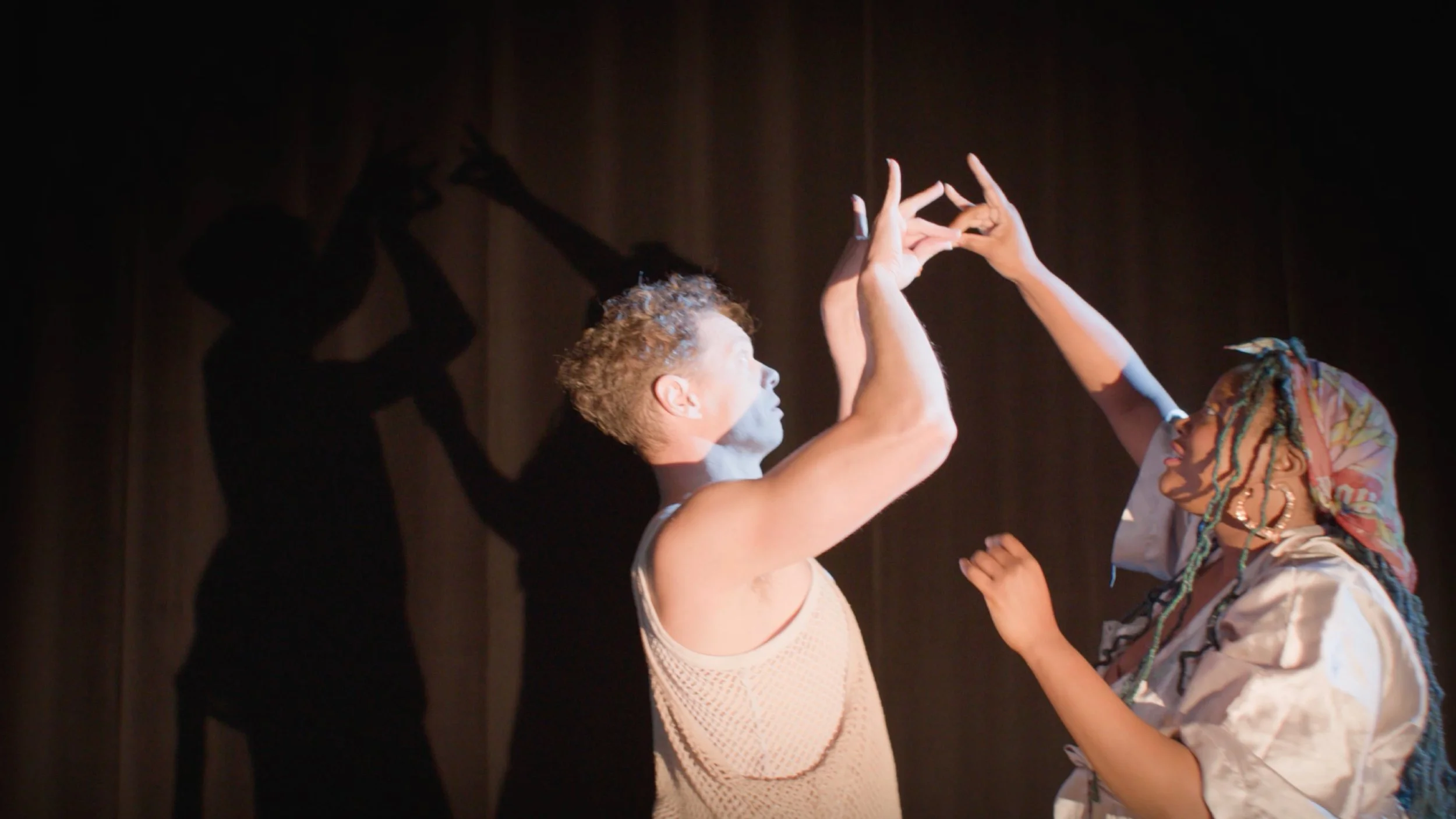 Two women are dancing and holding hands on a dark stage. The woman on the left has short curly hair and light skin, while the woman on the right has multicolored braided hair and darker skin.