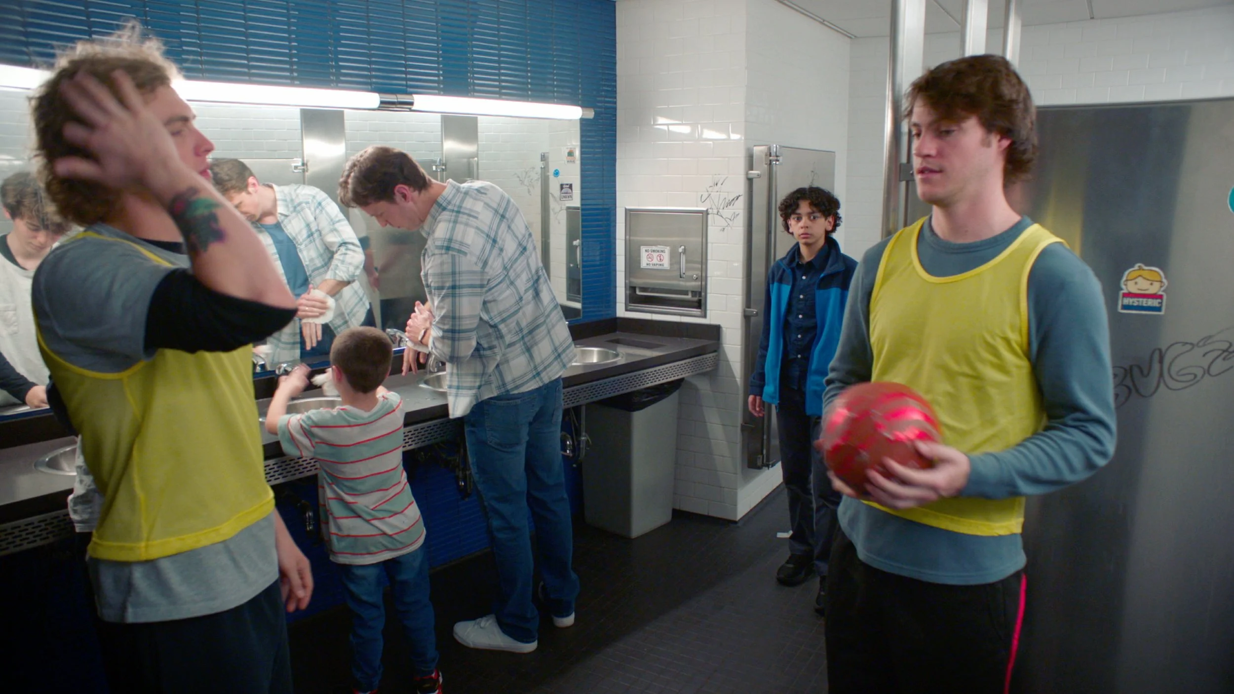 Group of young people in a restroom, some wearing yellow vests, engaging with a small child and holding a red ball, with a few others standing and talking.