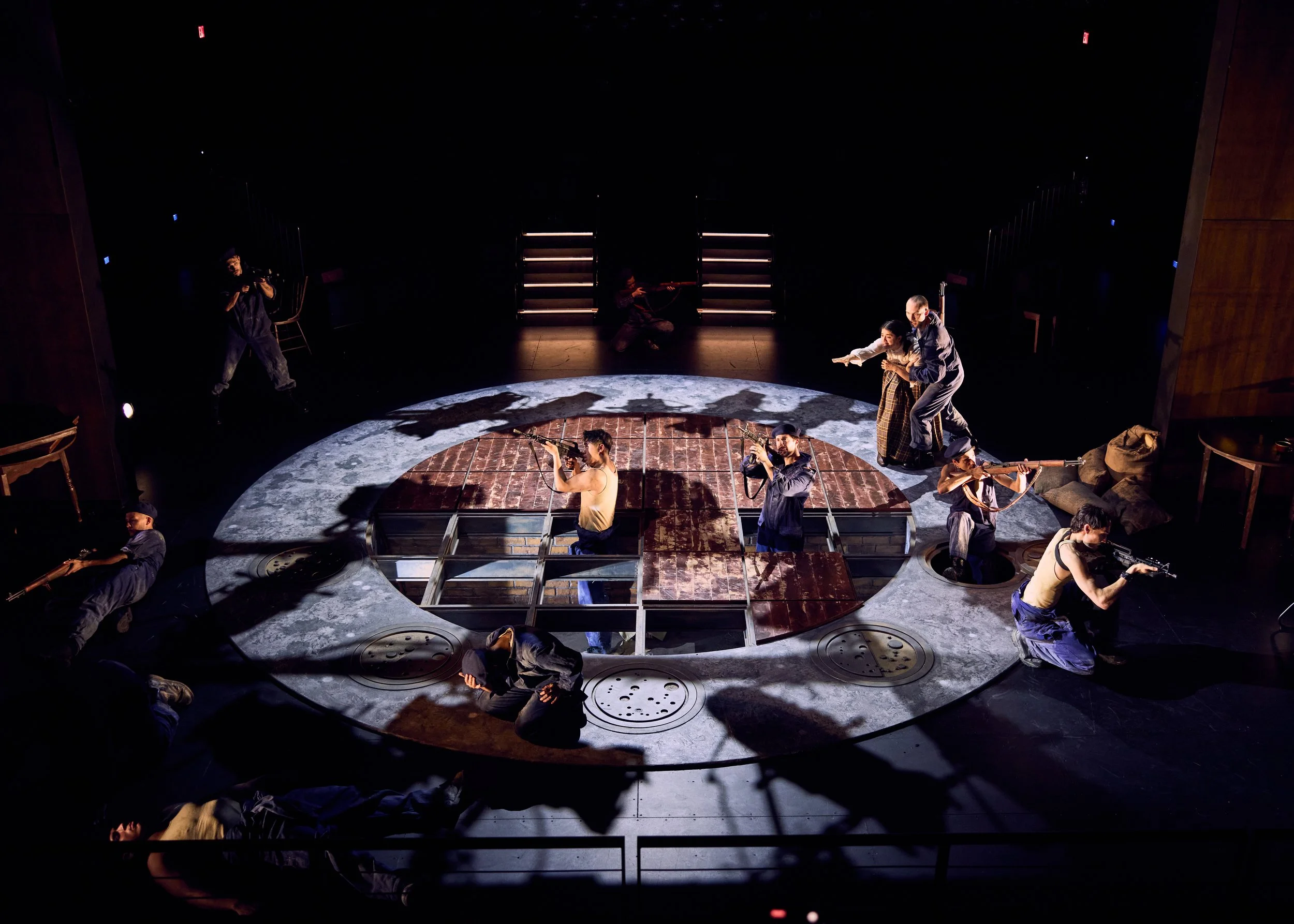 The image shows a theatrical stage scene with several actors in costume. The stage features a circular design with a brick floor section in the center, surrounded by a gray, moon-like surface. Actors are holding rifles, and some are crouching or knee