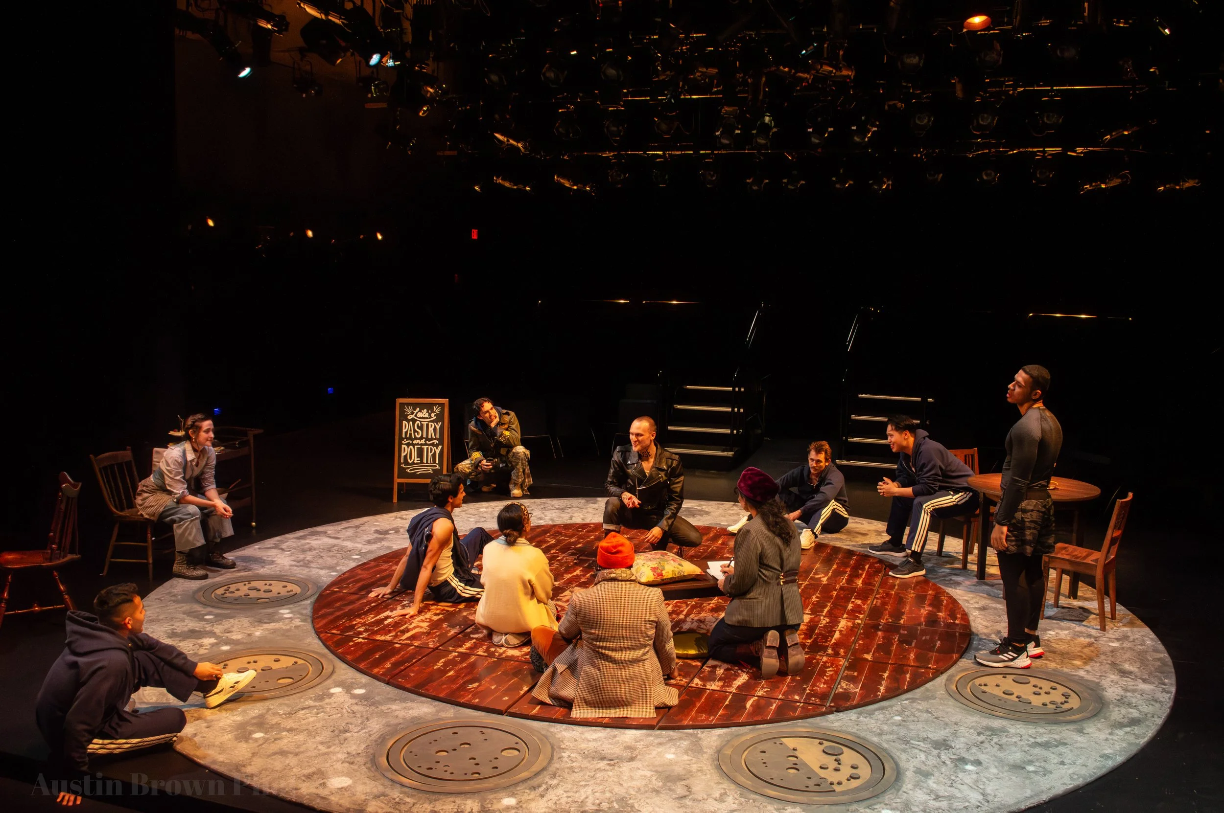 A stage set with actors and performers preparing for a theatrical production. The stage has a circular platform with some actors sitting and standing around it, and chairs and tables scattered nearby. There is a blackboard sign that reads 'Pastry and