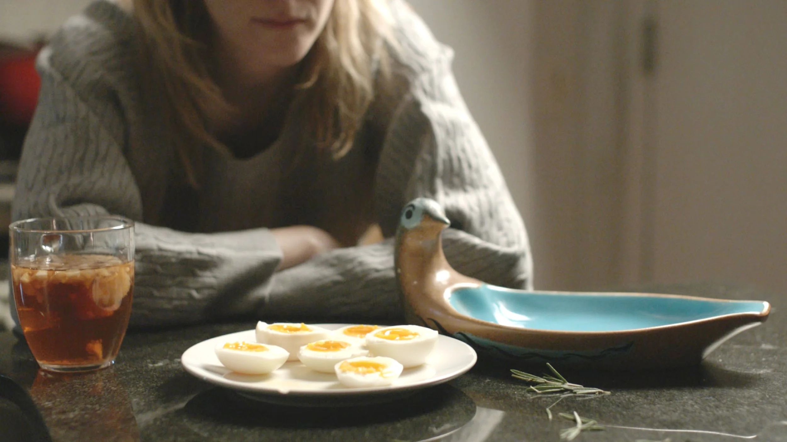 A woman sitting at a table with a glass of iced tea, a plate of halved soft-boiled eggs, a blue and brown ceramic dish shaped like a bird, and a sprig of rosemary on the table.