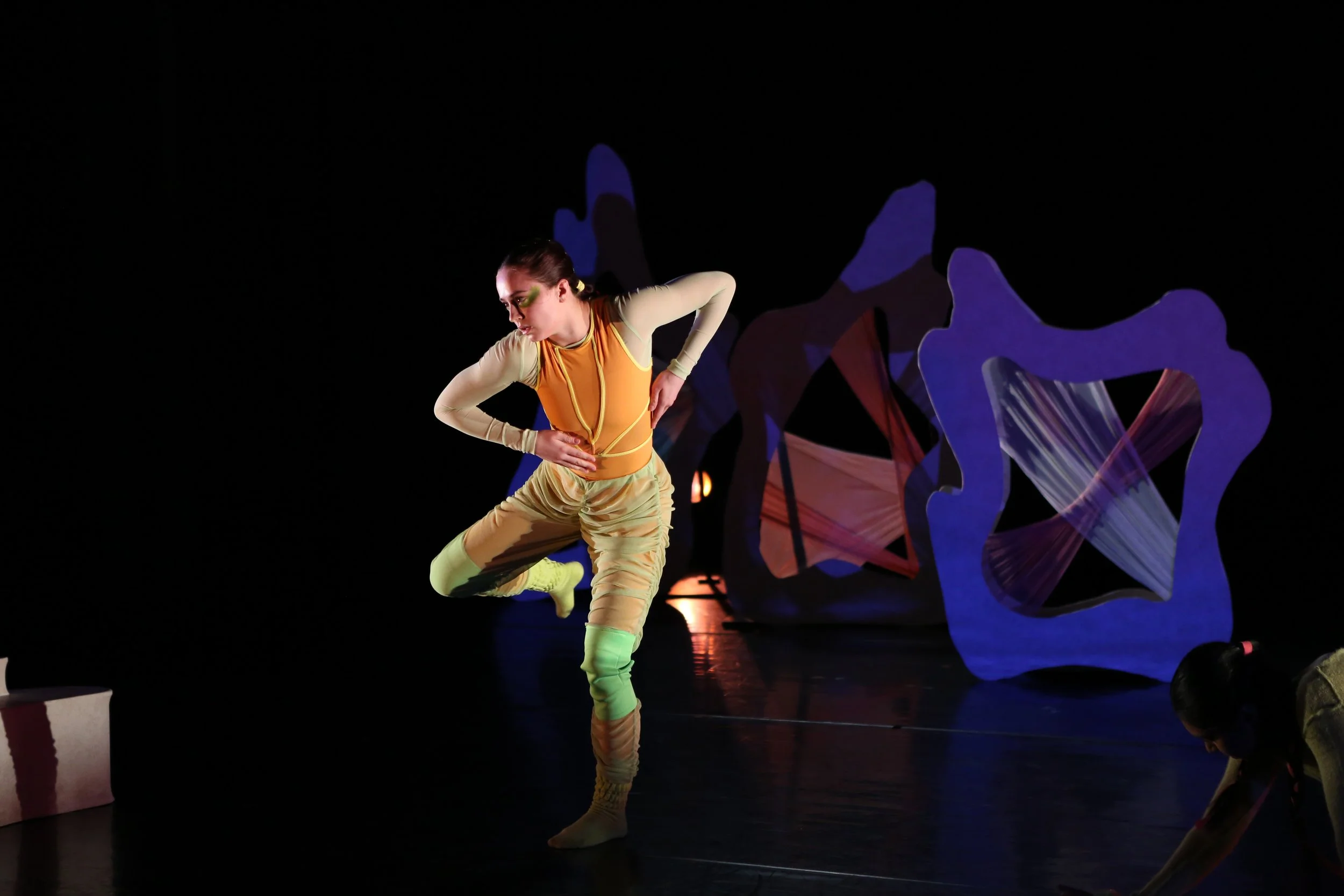 A female performer in colorful costume with face paint poses on stage, with abstract stage decorations illuminated behind her.