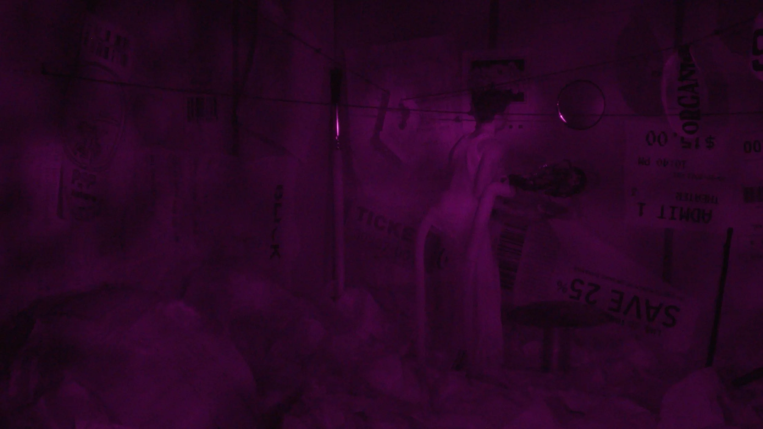 A dark, purple-tinted image of a person in a room with various signs and posters on the wall, some of which are partially visible and unclear.
