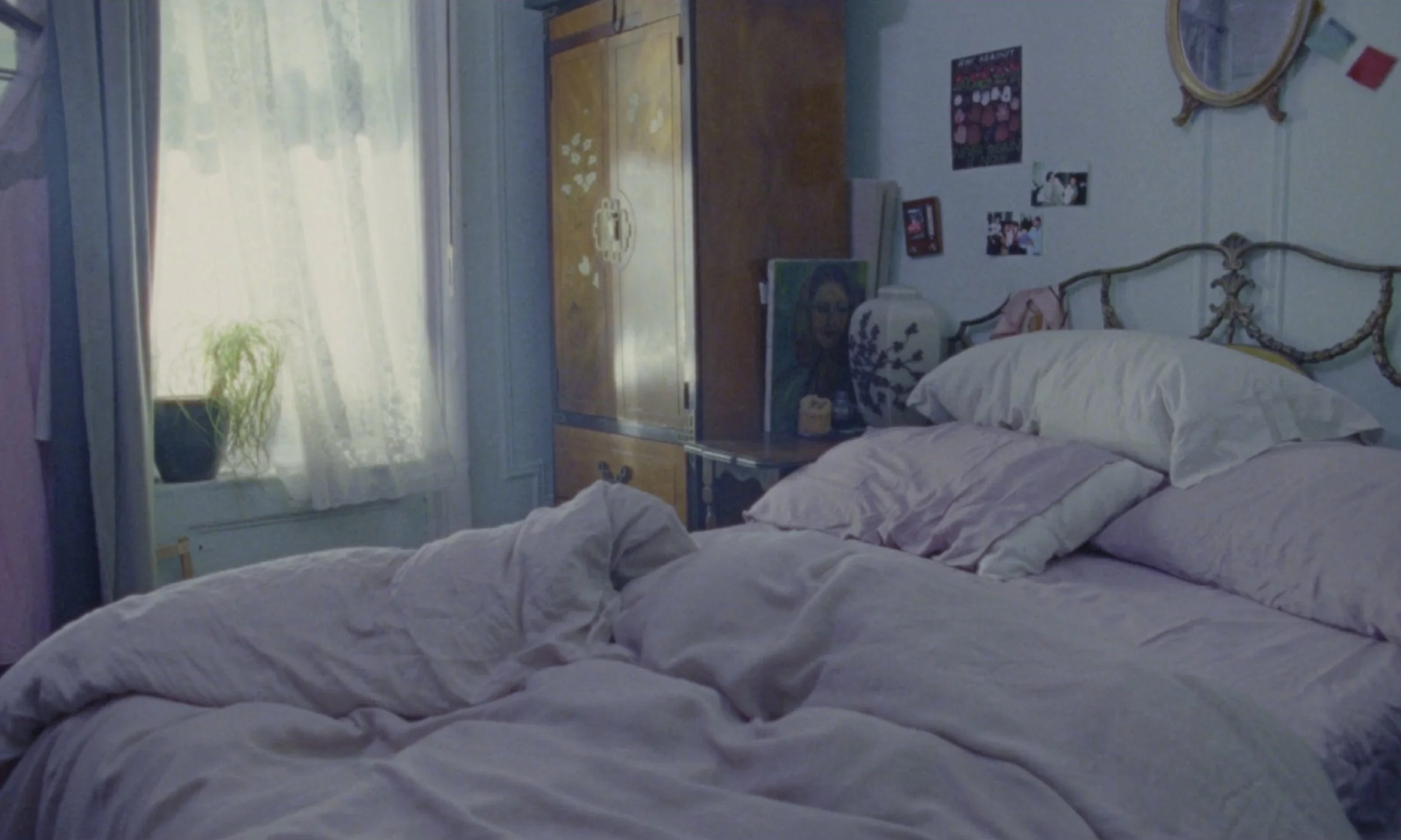 A bedroom scene with a bed that has unmade white bedding. There is a window with sheer curtains allowing sunlight to fill the room. A wooden dresser, some wall art, and photos are visible on the wall behind the bed. A potted plant is on the windowsil