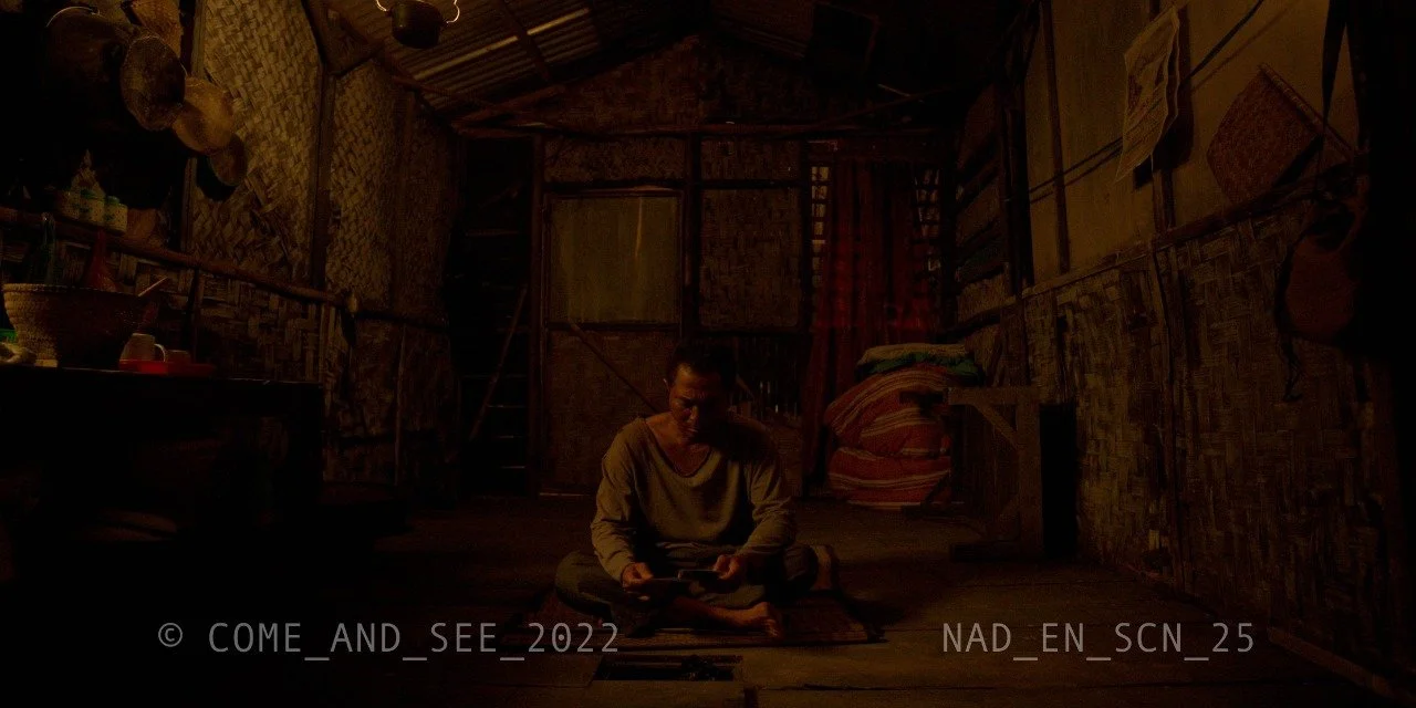 A person sitting on the floor inside a dimly lit rustic room, looking at a tablet or phone, surrounded by simple furniture and woven walls.
