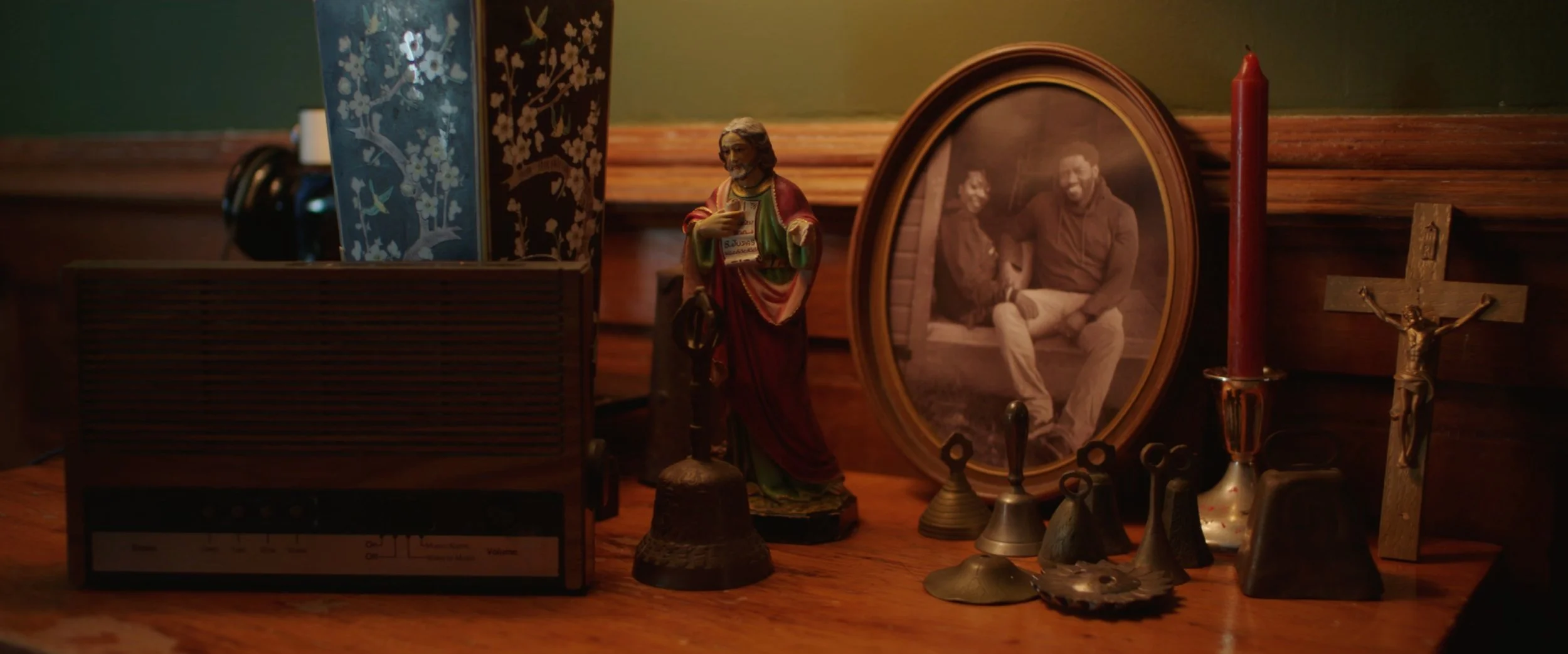A wooden table with religious and vintage items, including a crucifix, candle, religious figurine, and a framed black-and-white photo of two people sitting together, with a green wall and wooden trim in the background.