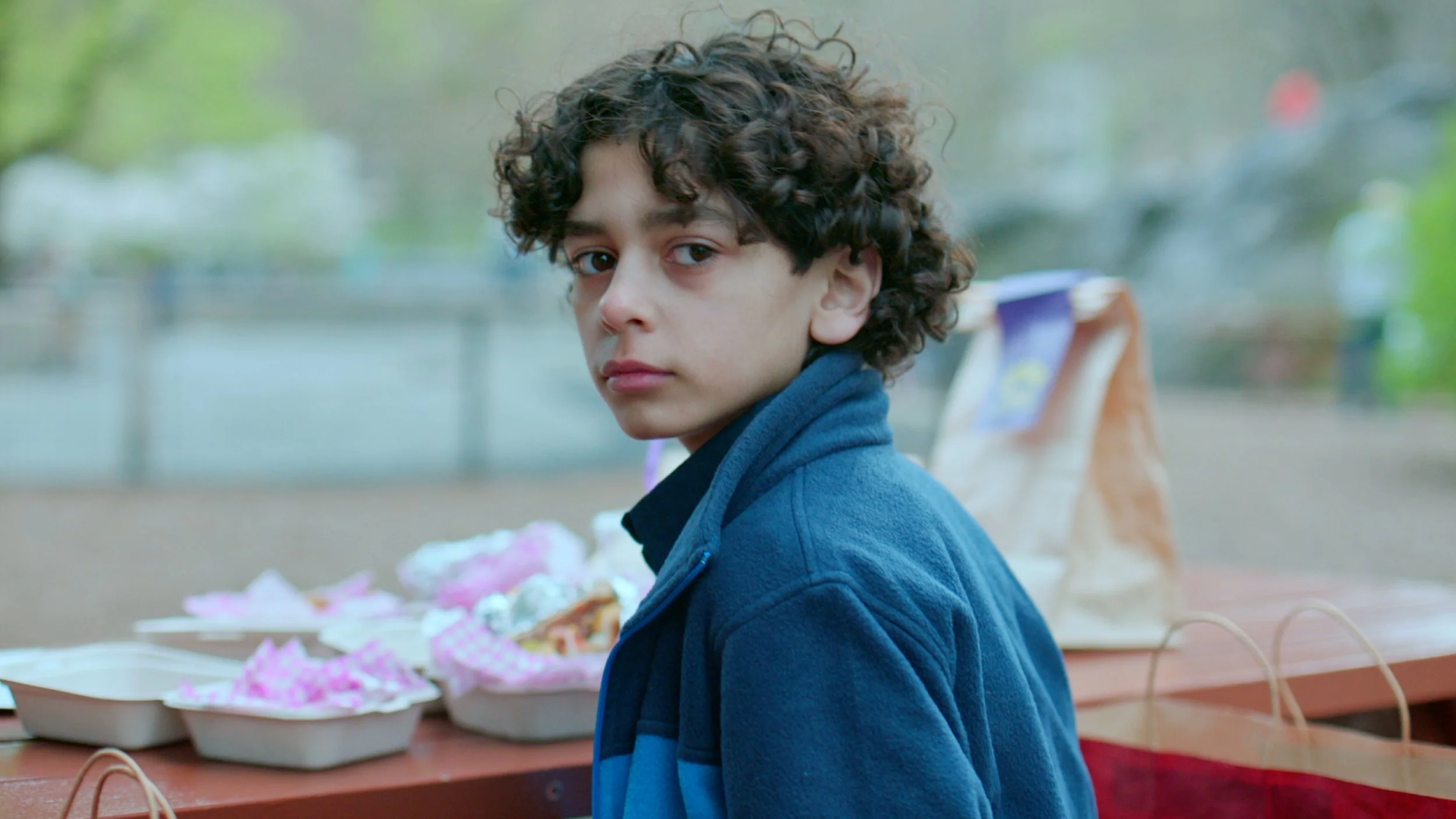 A young boy with curly dark hair and light skin is sitting in front of a table with colorful wrapped candies and snacks, looking over his shoulder.