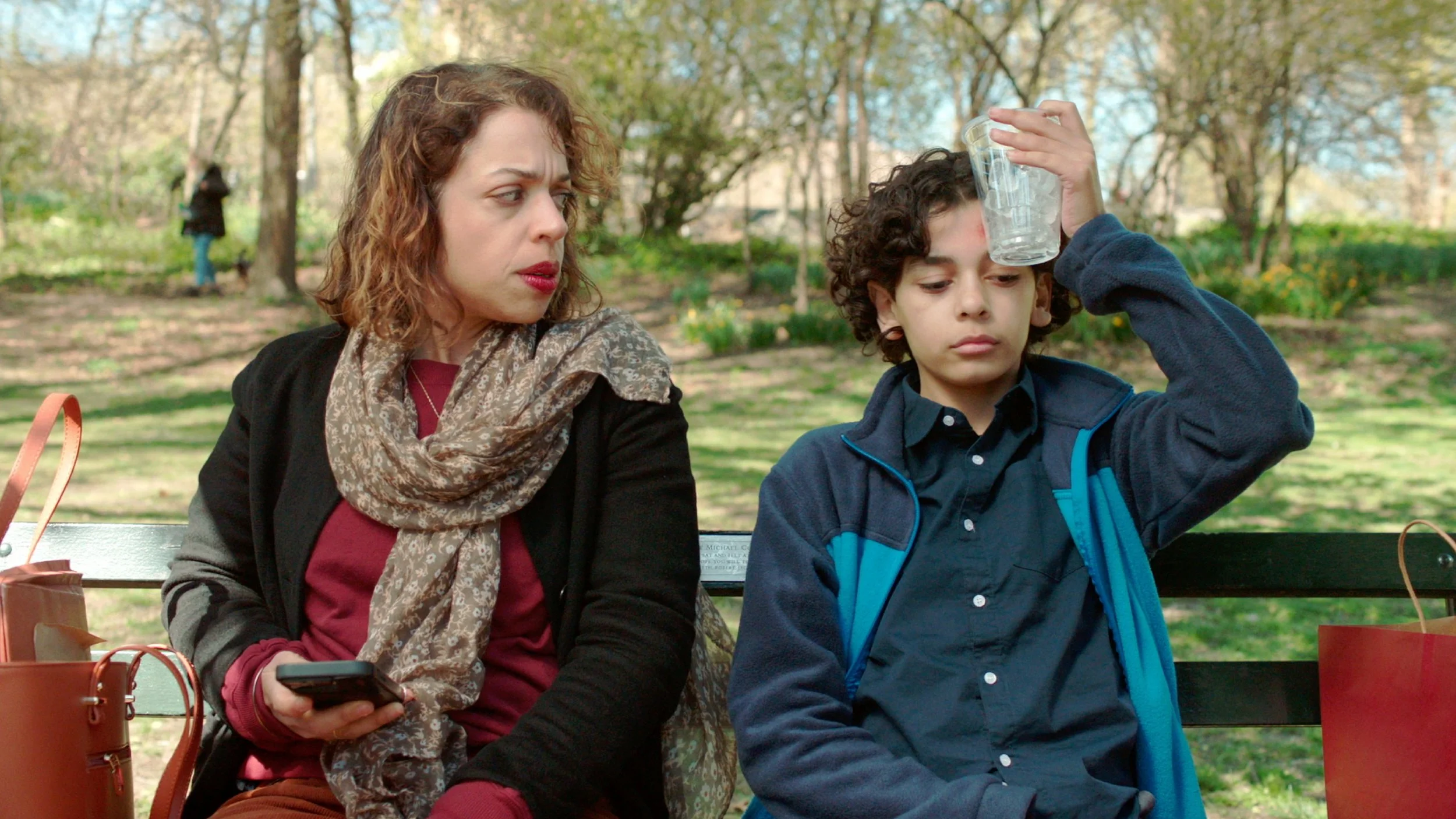 A woman and a boy sitting on a park bench. The woman is looking at the boy, who is holding a clear plastic cup on his forehead. The woman is holding a smartphone in her hand. There are trees and other people in the background.
