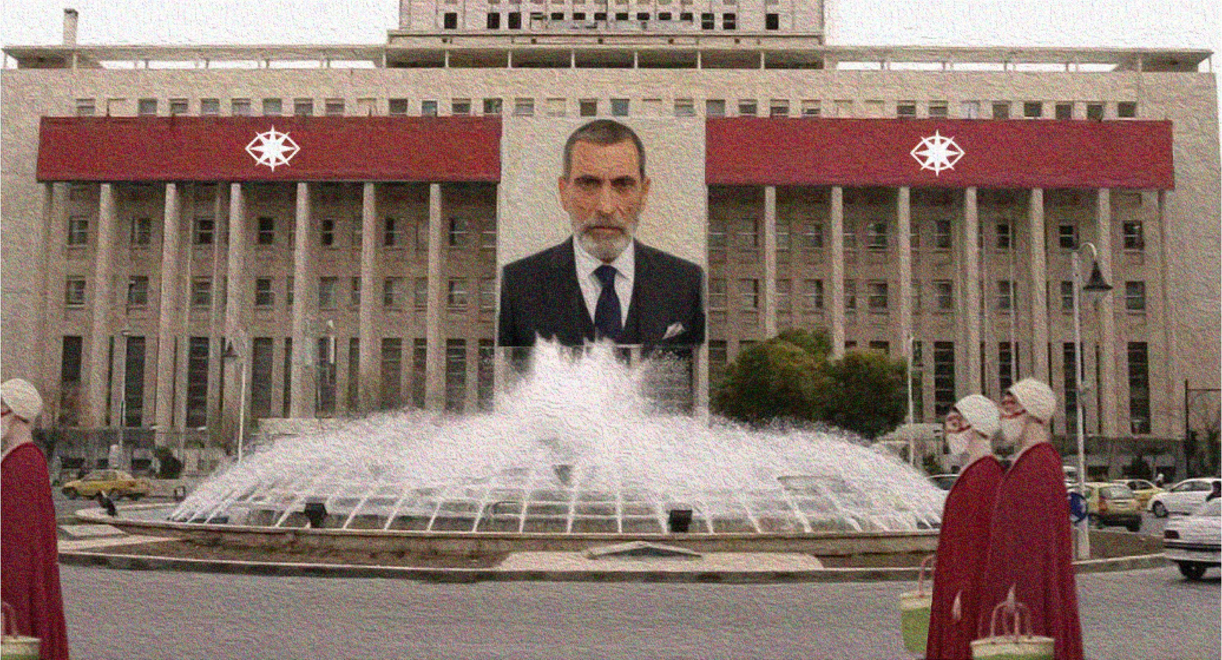 A large building with red banners featuring a white emblem on the balcony area. In front, there is a fountain with water spraying upward. Some people are seated nearby, and cars are parked on the street.