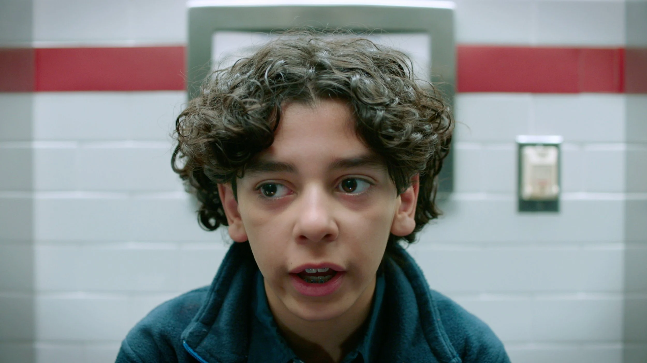 Close-up of a young boy with curly hair and freckles, sitting against a background of white and red tiles, looking intently at the camera.