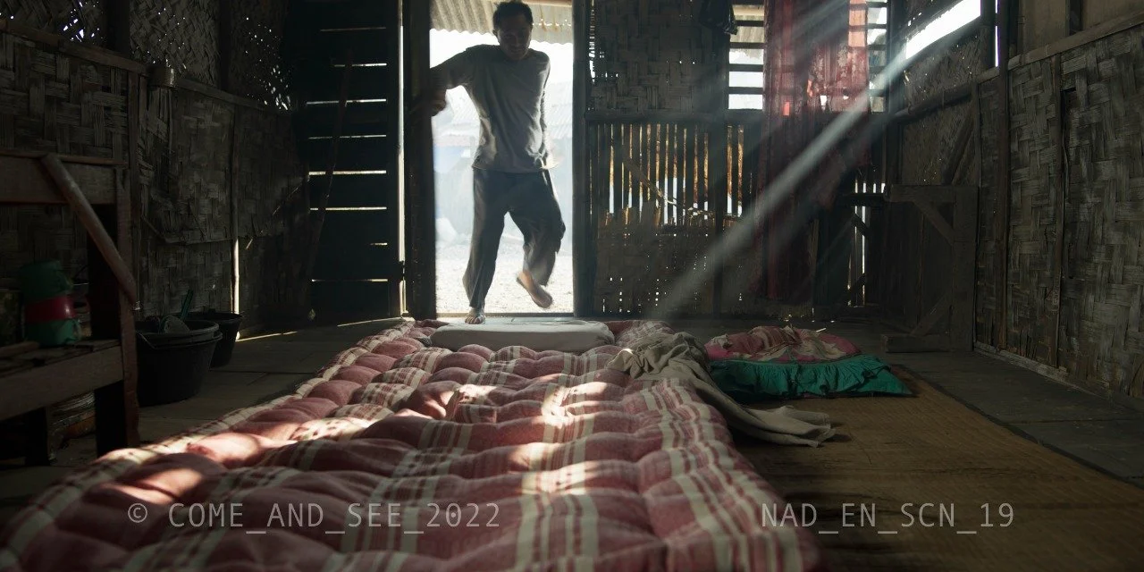 Silhouette of a man entering a wooden hut through the doorway, sunlight streaming inside, with bedding and pillows on the floor.