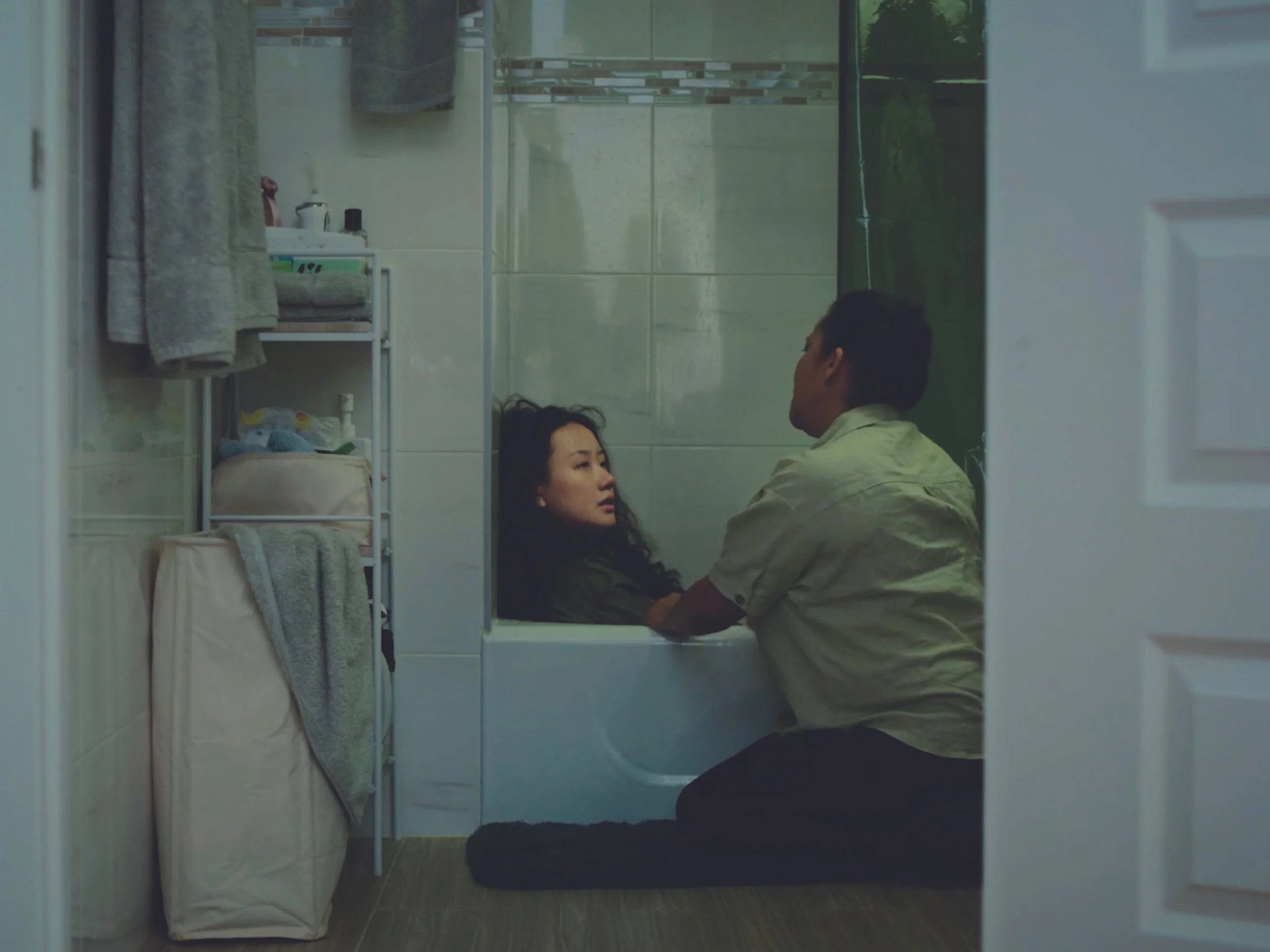 A woman lying in a hospital bed, with two men kneeling beside her, in a small bathroom or medical room.