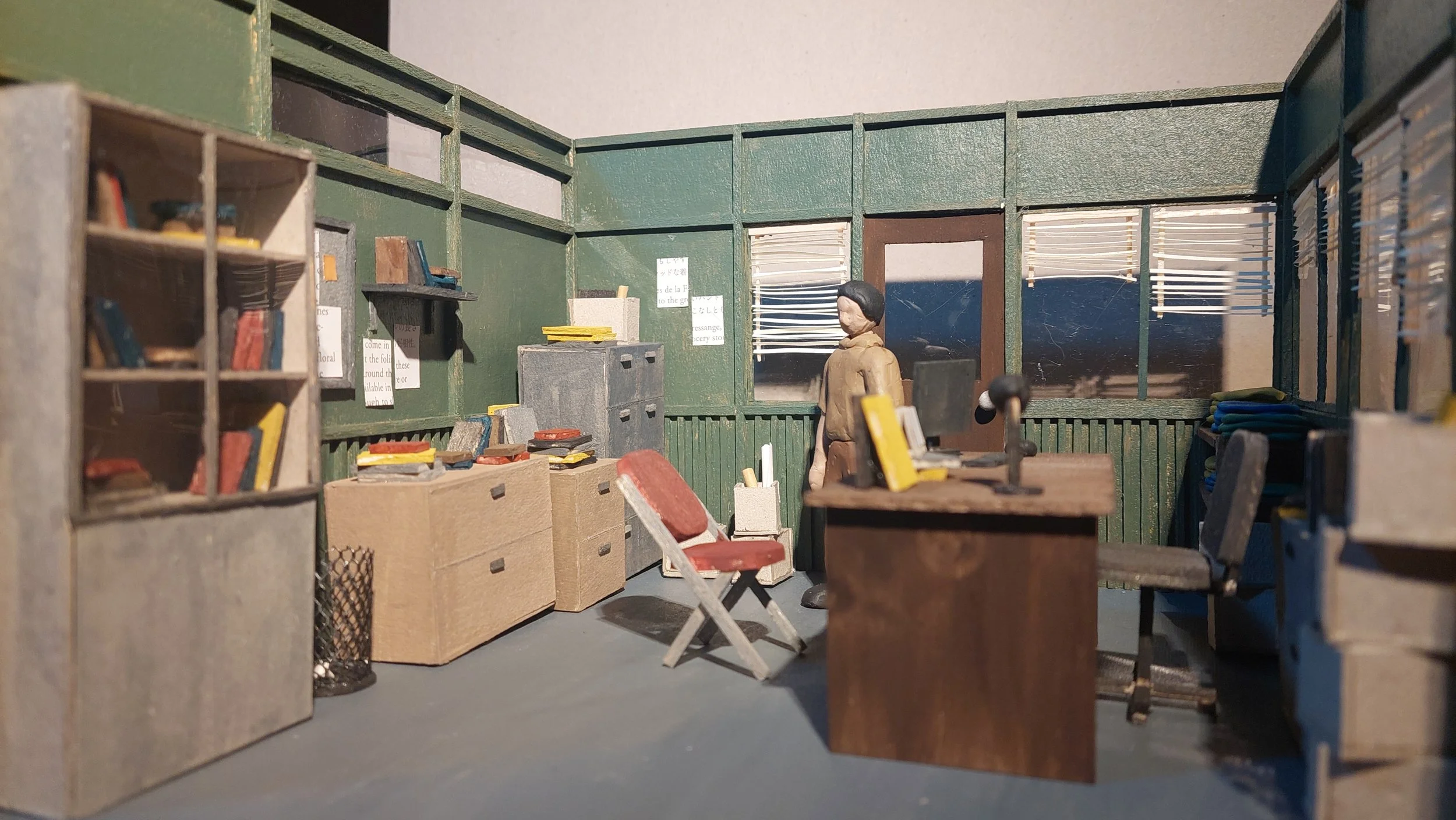 Miniature model of a library room with shelves filled with books, a desk, and a figure of a person standing near the window.