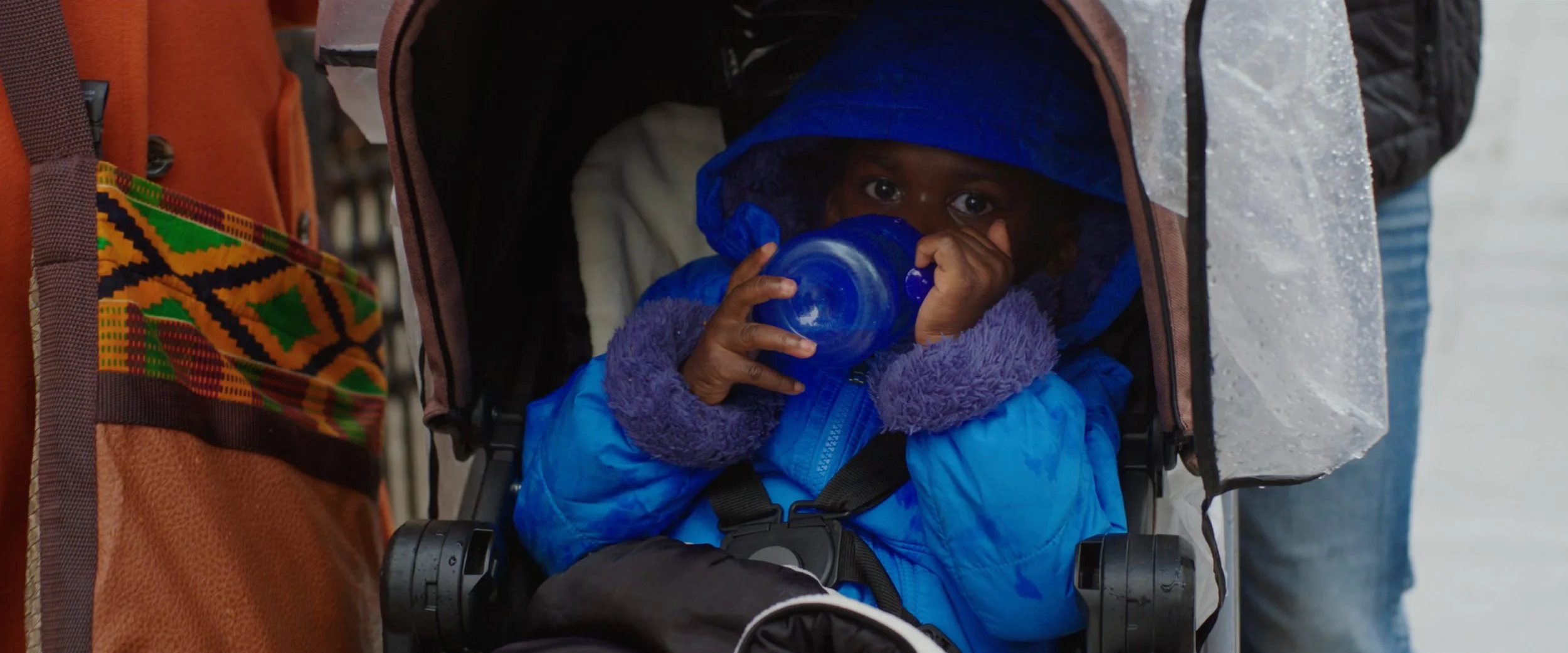 A young child dressed in a blue jacket and purple fleece kept warm, sitting in a stroller and drinking from a blue bottle, in a cold environment with snow and ice, surrounded by people.