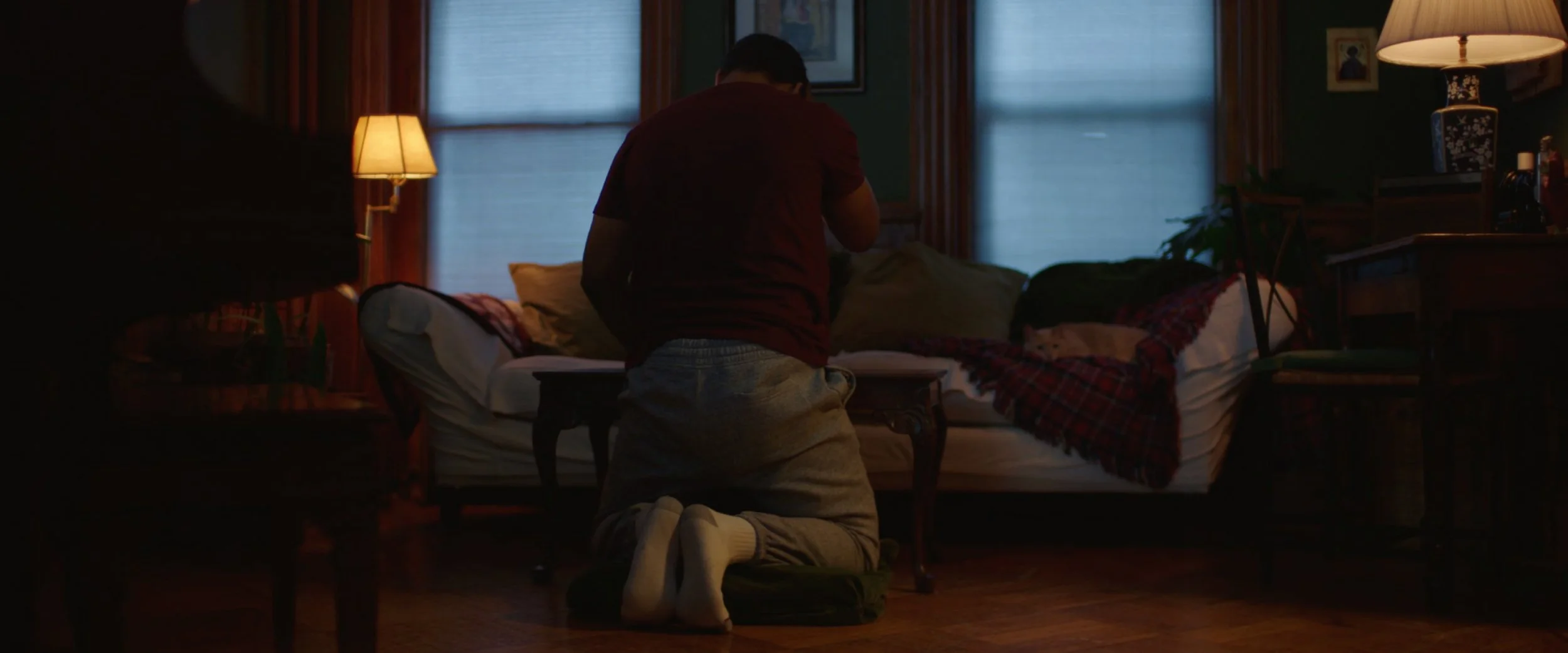 A person kneeling on the floor in front of a bed in a dimly lit room, with two windows, lamps, and various furniture and decorations.