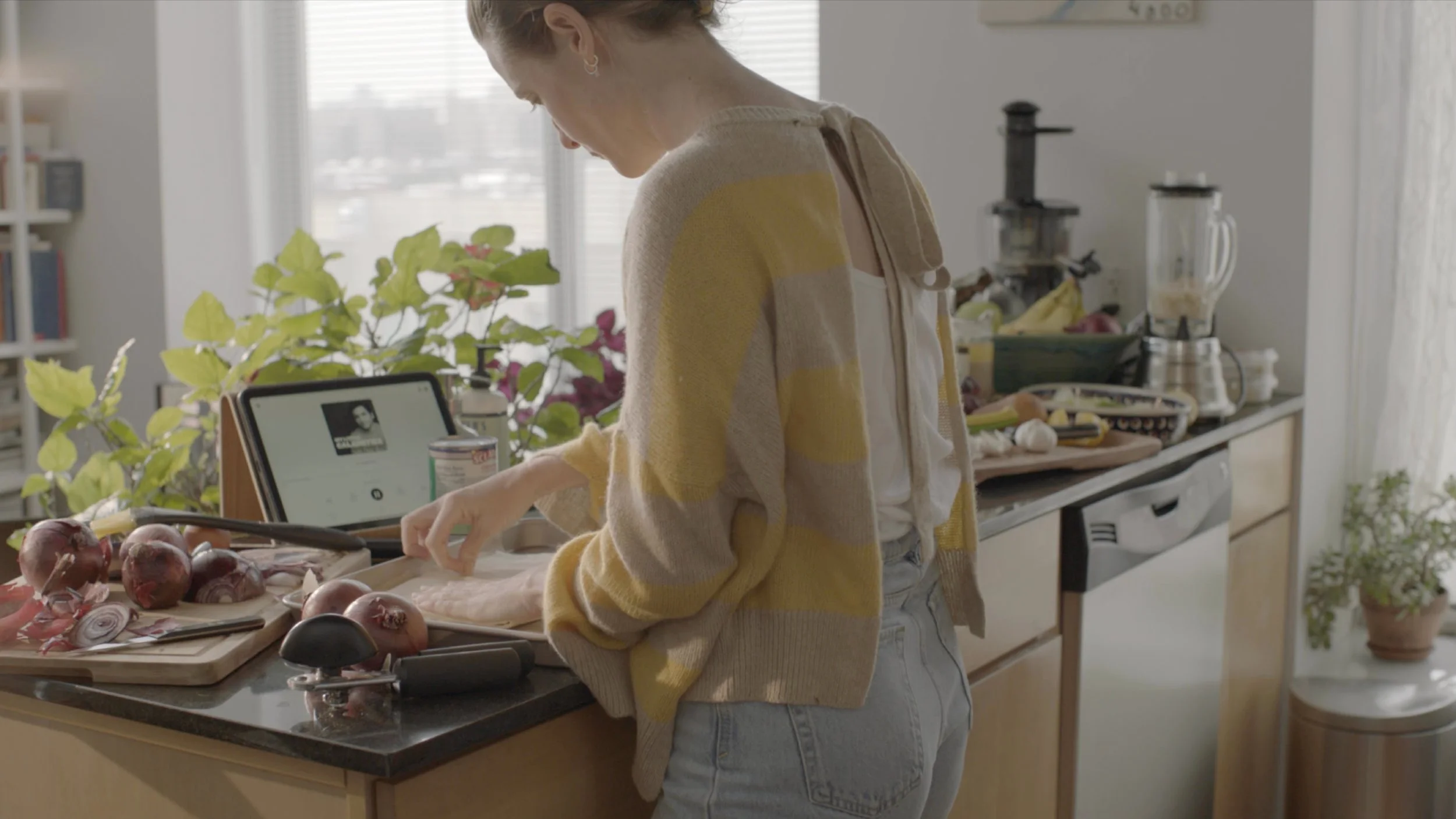 A woman cooking in a kitchen with onions on a cutting board, green plants, a tablet, and various kitchen appliances on the counter.