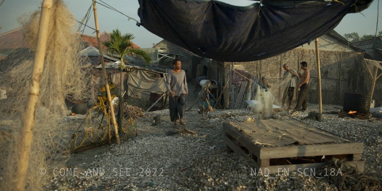 Scene of a makeshift outdoor workspace with several men working under a large black tarp. The area is cluttered with various materials, debris, and a pallet on the gravel ground. Some men are handling a plastic sheet or tarp.