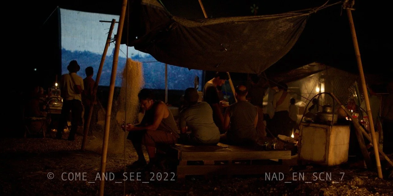 People sitting and standing inside a tent made of wooden poles and fabric at night, with a large projection screen visible in the background and a traditional stove or cooking area on the right.