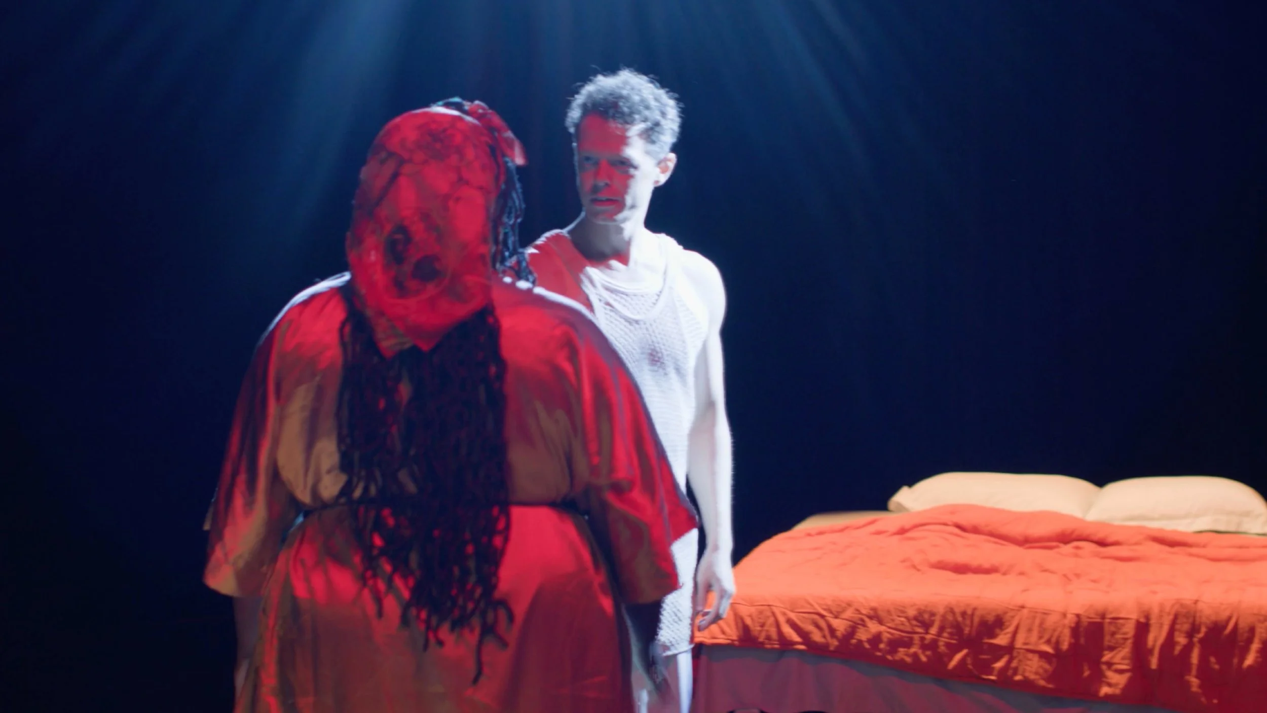 Two people standing near a bed with an orange blanket. One person is wearing a red hoodie with black lace details on the sleeves, and the other person is wearing a white sleeveless top. The scene is dark with a spotlight on them.