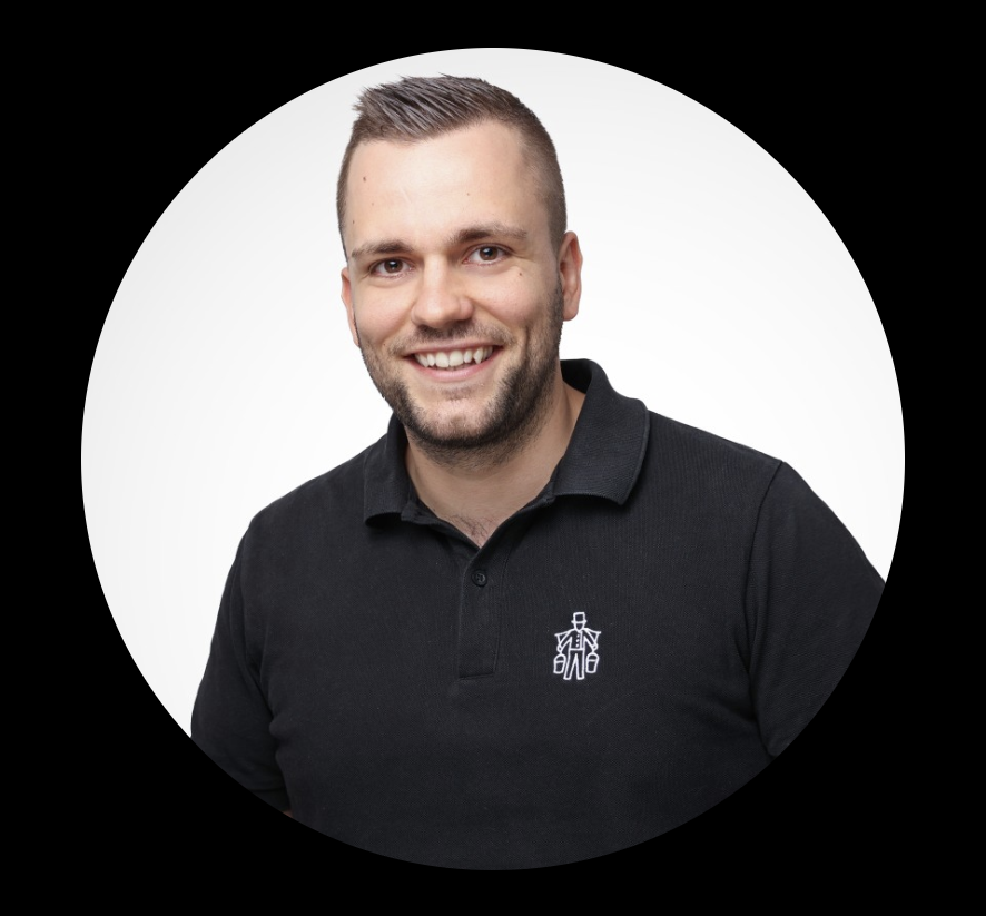 Ein lachender Mann mit heller Haut, braune Haare und Bart, trägt ein schwarzes Polo-Shirt mit einem weißen Logo auf der linken Brustseite, vor einem weißen und schwarzen Hintergrund.