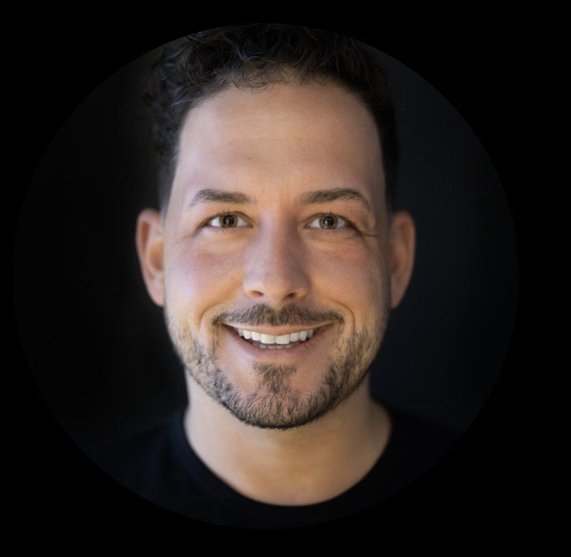 Nahaufnahme eines lachenden Mannes mit dunklem Haar, Vollbart und hellen Augen vor schwarzem Hintergrund.