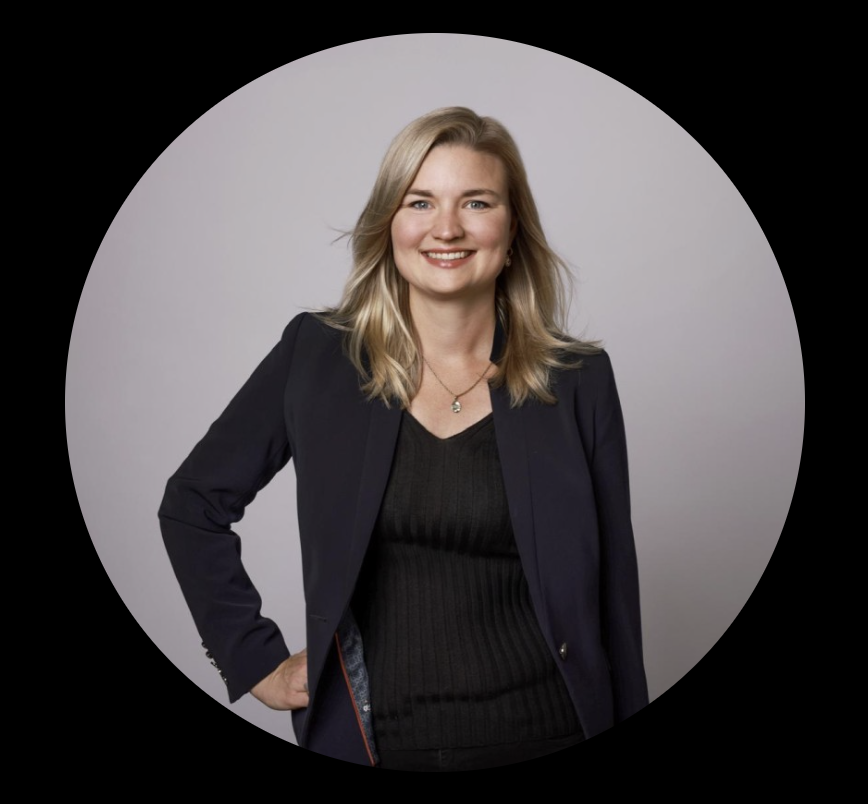 Ein lächelnder blonde Frau in dunkler Jacke und schwarzem Top vor grauem Hintergrund.