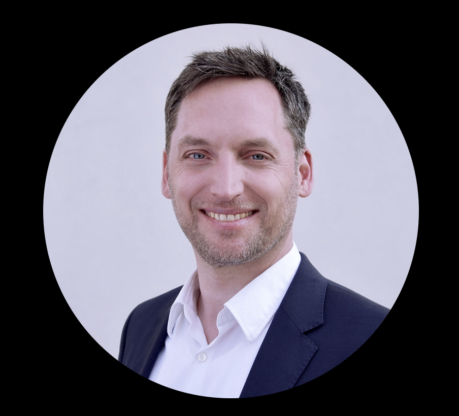 Porträt eines lächelnden Mannes mit braunem Haar, blauen Augen, in einem weißen Hemd und einem schwarzen Anzug. Hintergrund ist weiß und das Bild ist kreisförmig.