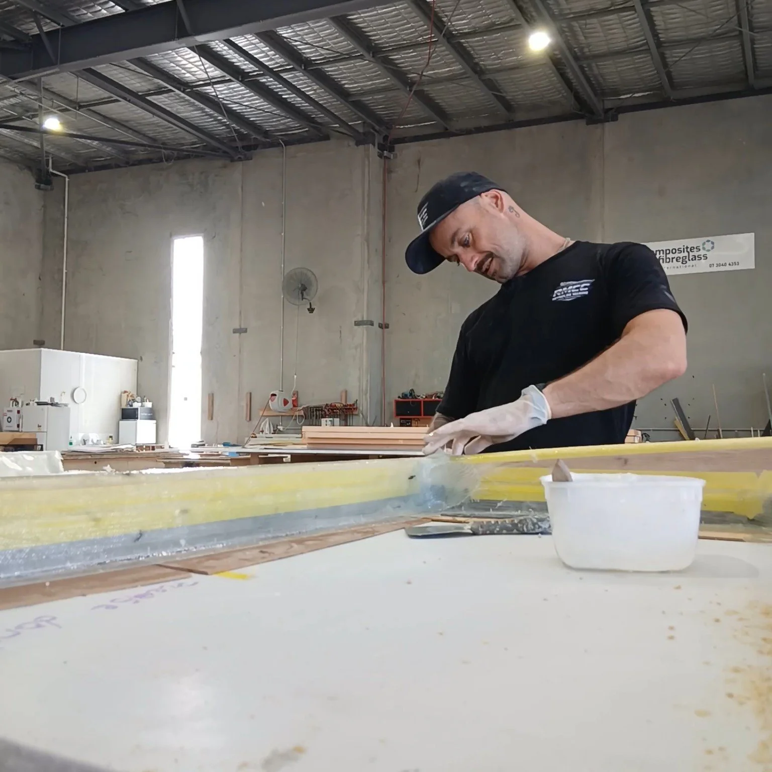 A man in a black hat and shirt is working in a workshop, focusing on a large yellow piece of material, with tools and materials around him.