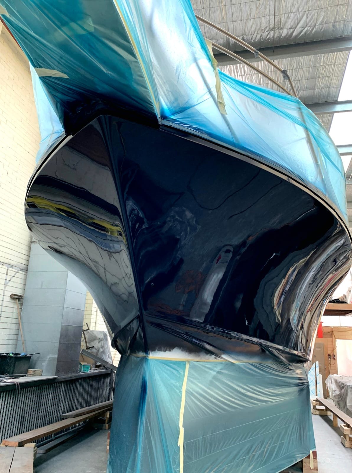 A large boat inside a workshop, with the hull painted black and the upper part covered with blue protective film.