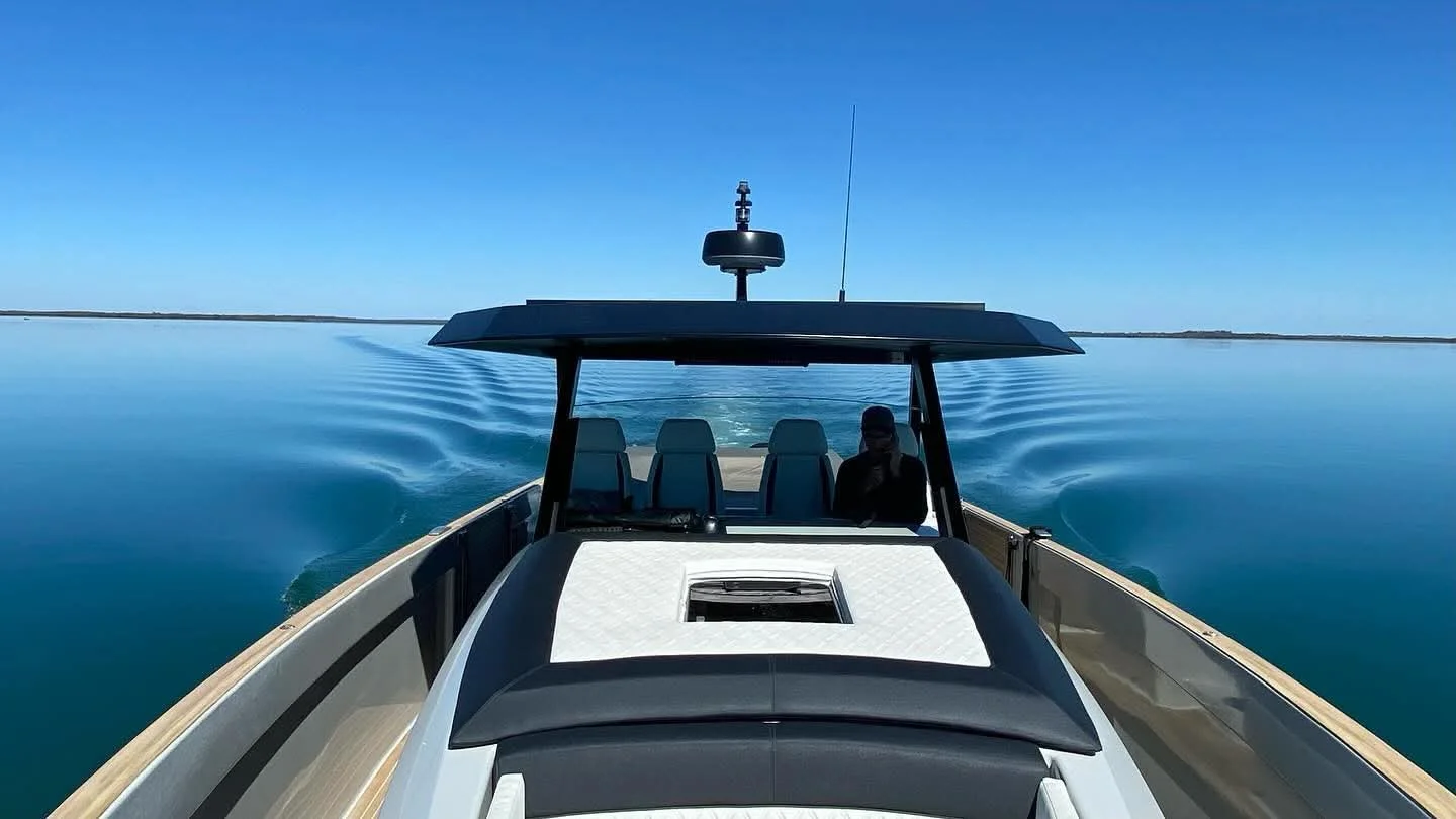 A modern boat on calm water under a clear blue sky, viewed from the front showing the boat's bow, a canopy with seats, and a person sitting on the right side.