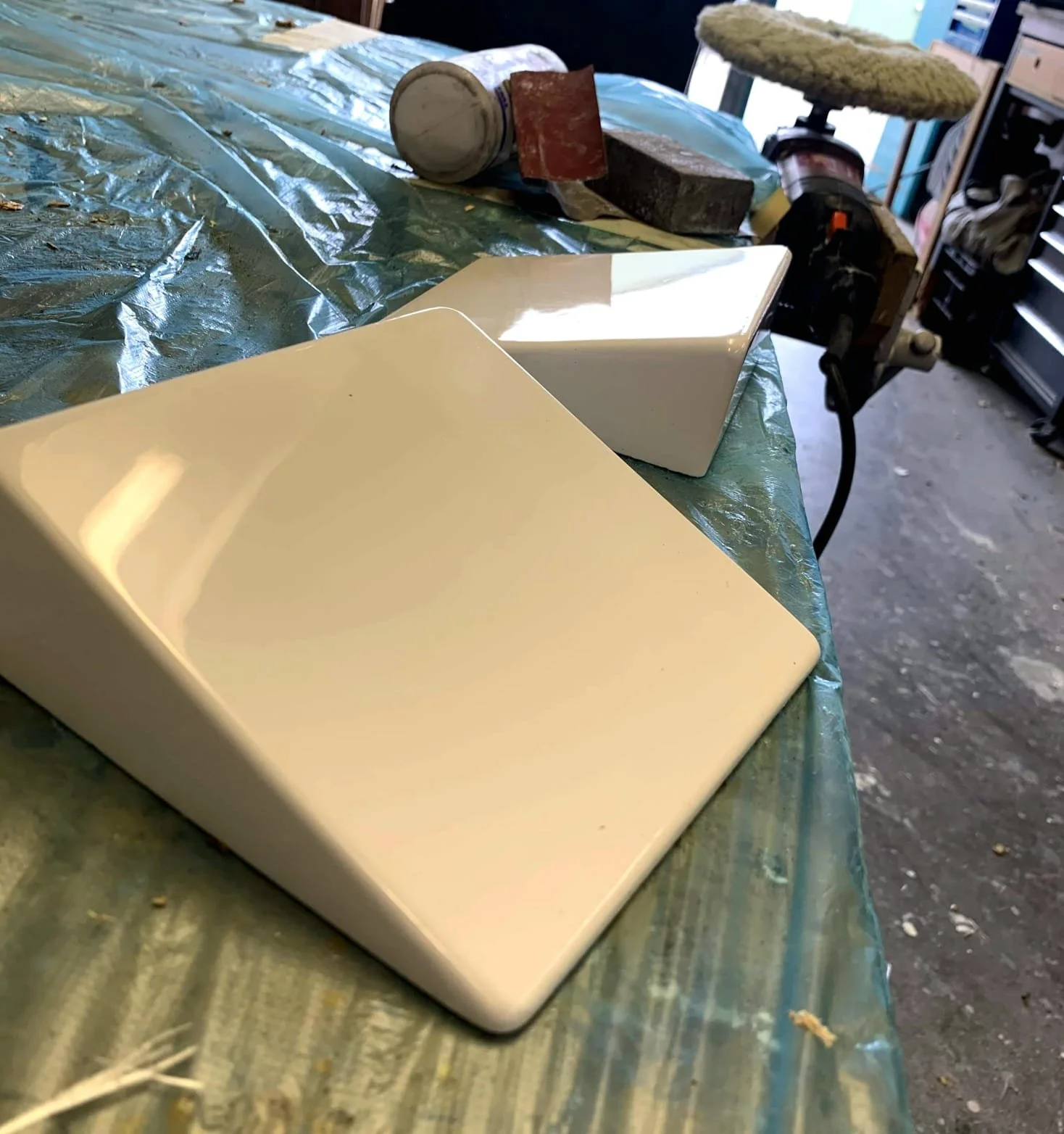 Two white rectangular objects, possibly sinks or containers, on a plastic-covered work surface in a workshop or garage. The background shows tools, sanding and polishing equipment, and an uneven floor.