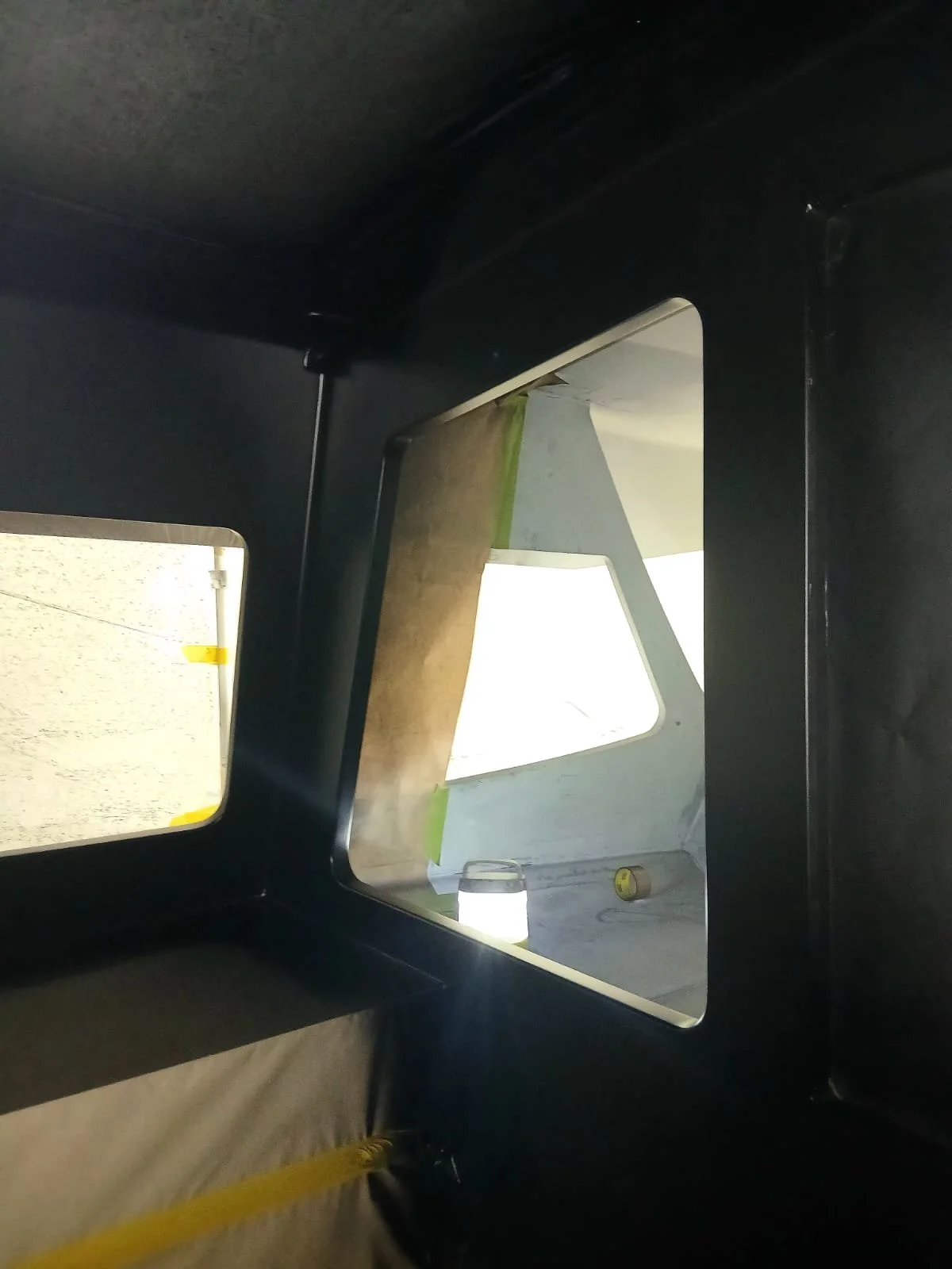 Interior of a dark vehicle, looking out the window at a light-colored structure with a window and some objects outside.