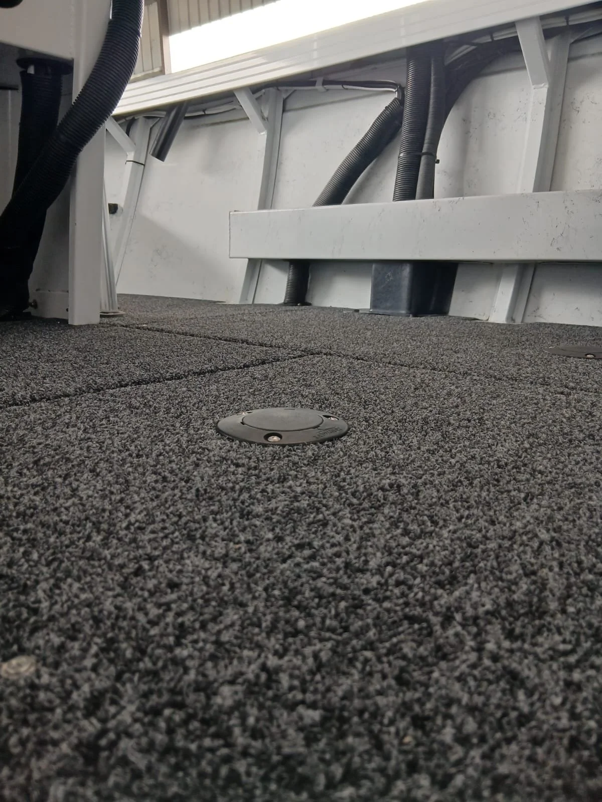 Close-up of a gray carpeted floor with a sealed circular floor access cover, under a structure with black flexible conduit and metal framing.