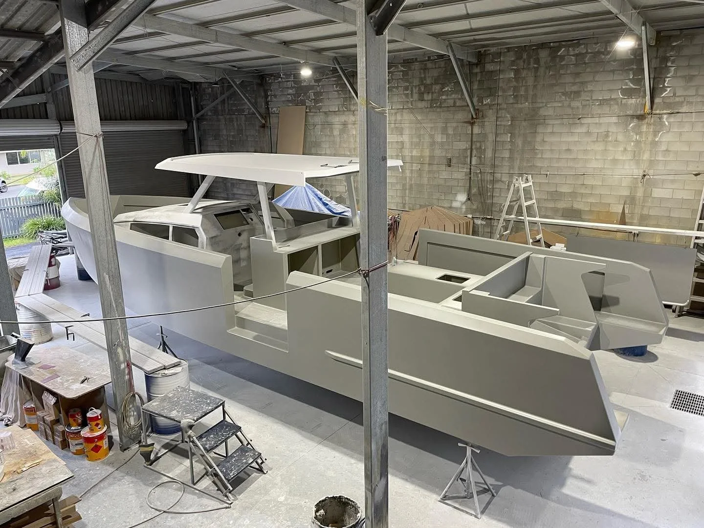 An unfinished boat model in a workshop with tools, ladders, and supplies around.