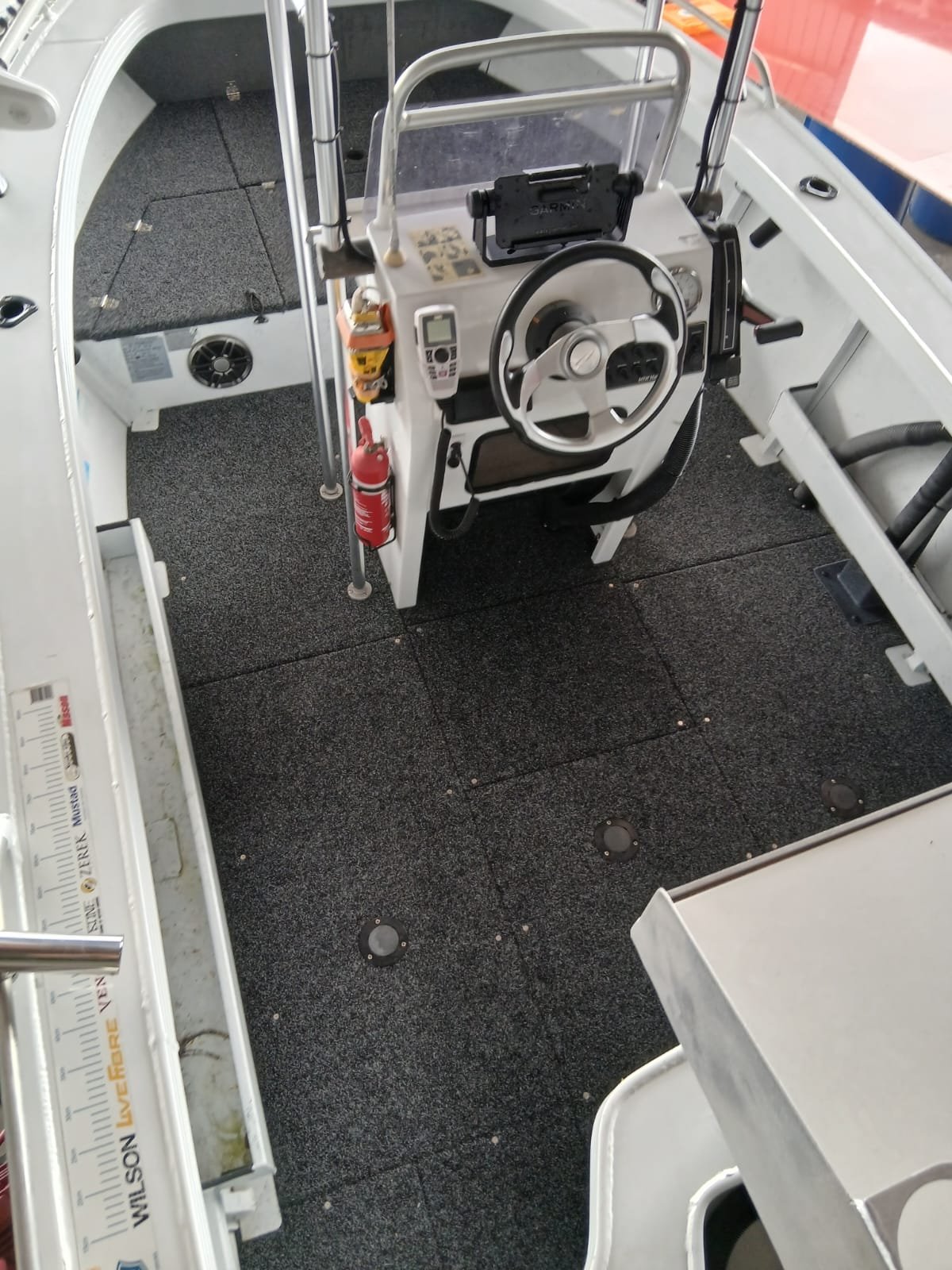 Interior of a boat cabin with a steering wheel, electronic device, fire extinguisher, and seating area, with a black textured floor.