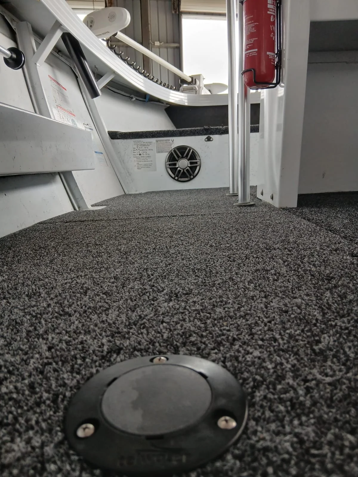 View from a low angle showing a boat interior with carpeted flooring, a fire extinguisher attached to a metal pole, a ventilation vent on the wall, and a window with natural light coming through.