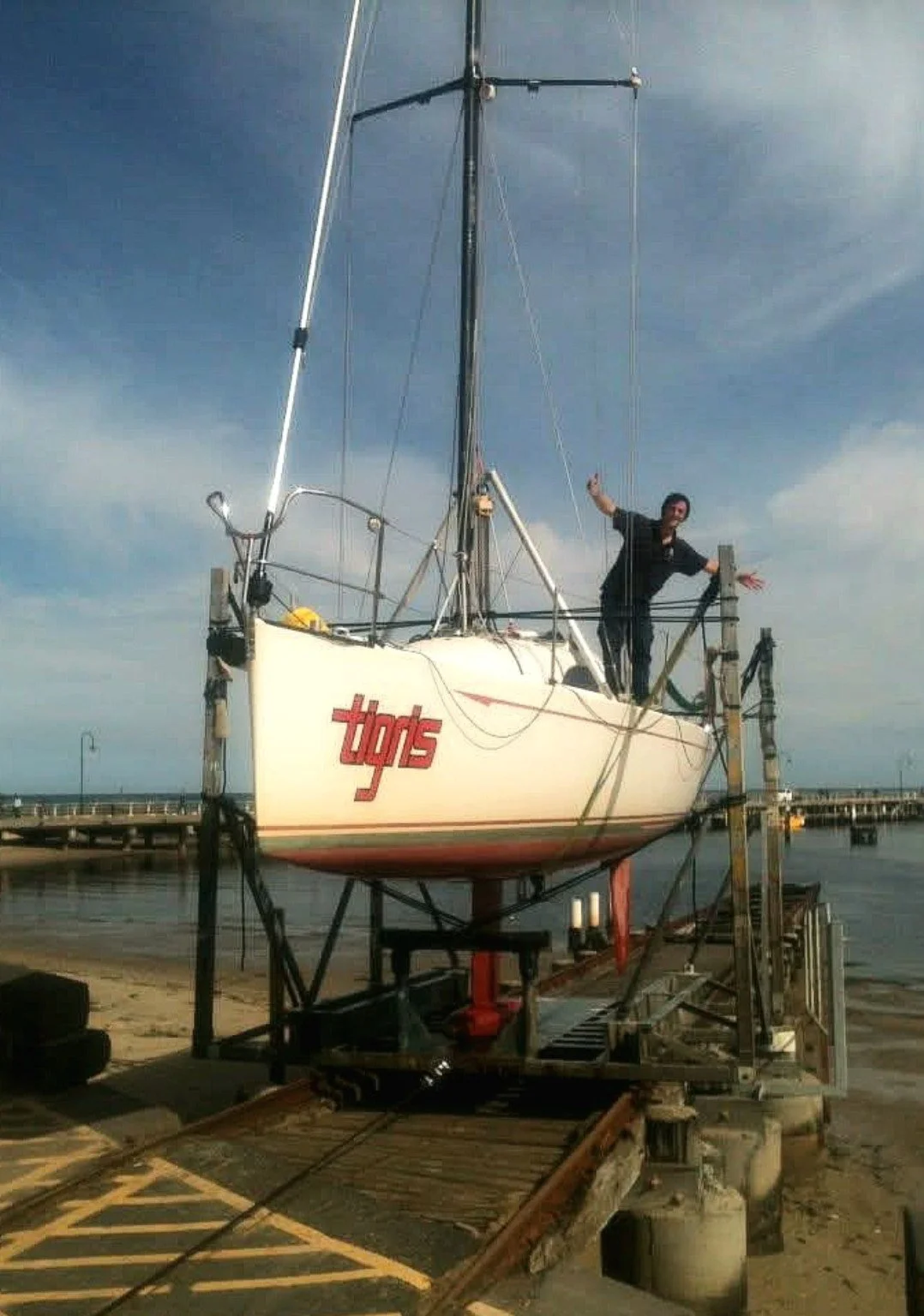 Person standing on a boat on a dry dock, waving and smiling at the camera. The boat has the word 'typos' written on its side.